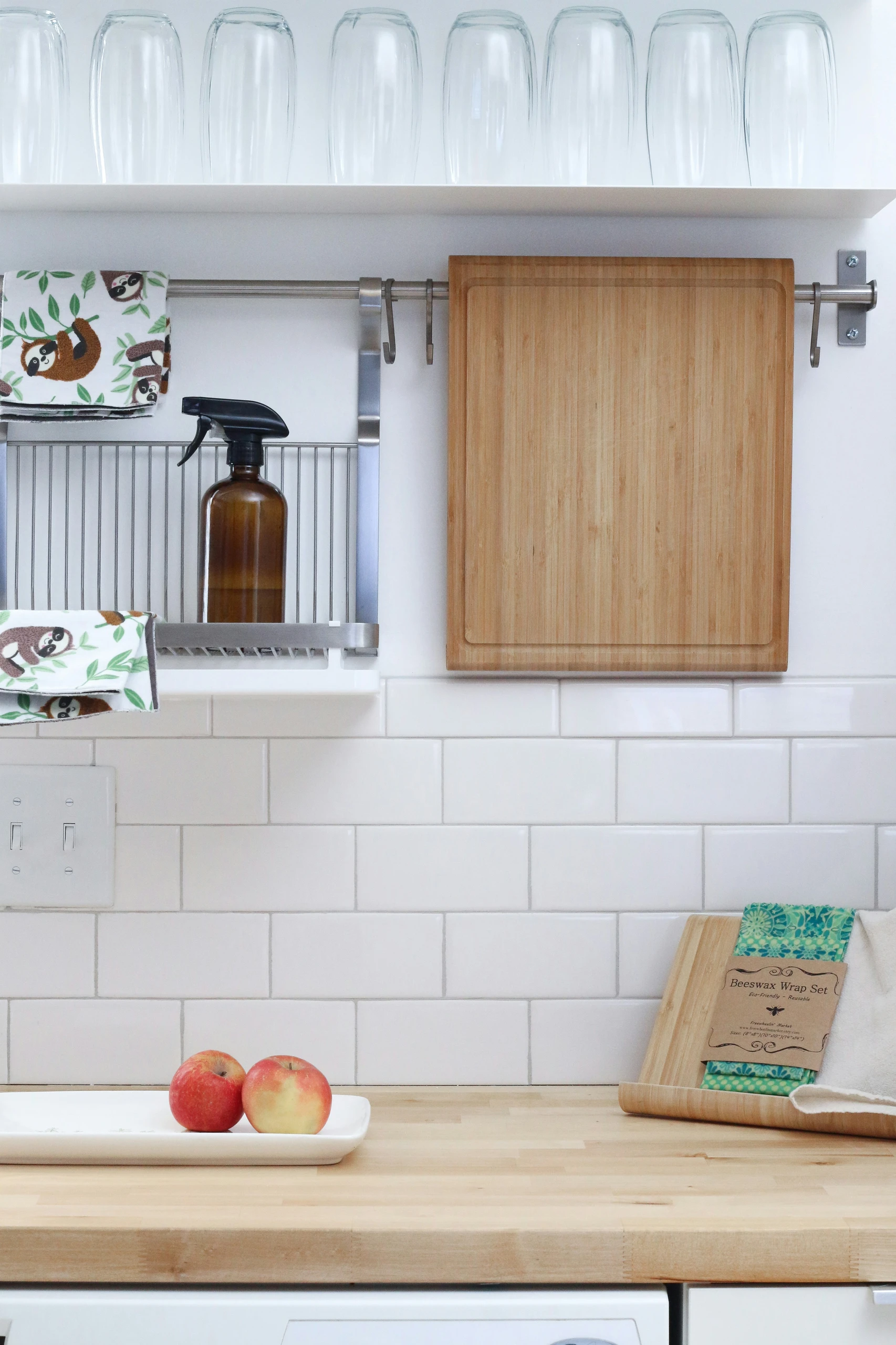 A bright and tidy kitchen scene with a wooden countertop holding two apples on a white tray and a wooden cutting board with beeswax wraps. Above the counter, a sleek metal ray is mounted to the wall, used to hang up your kitchen utensils on a ray, including a cutting board, patterned towels, and a brown glass spray bottle for easy access. An inspiring example of kitchen counter organization, blending practicality with a clean, modern style.