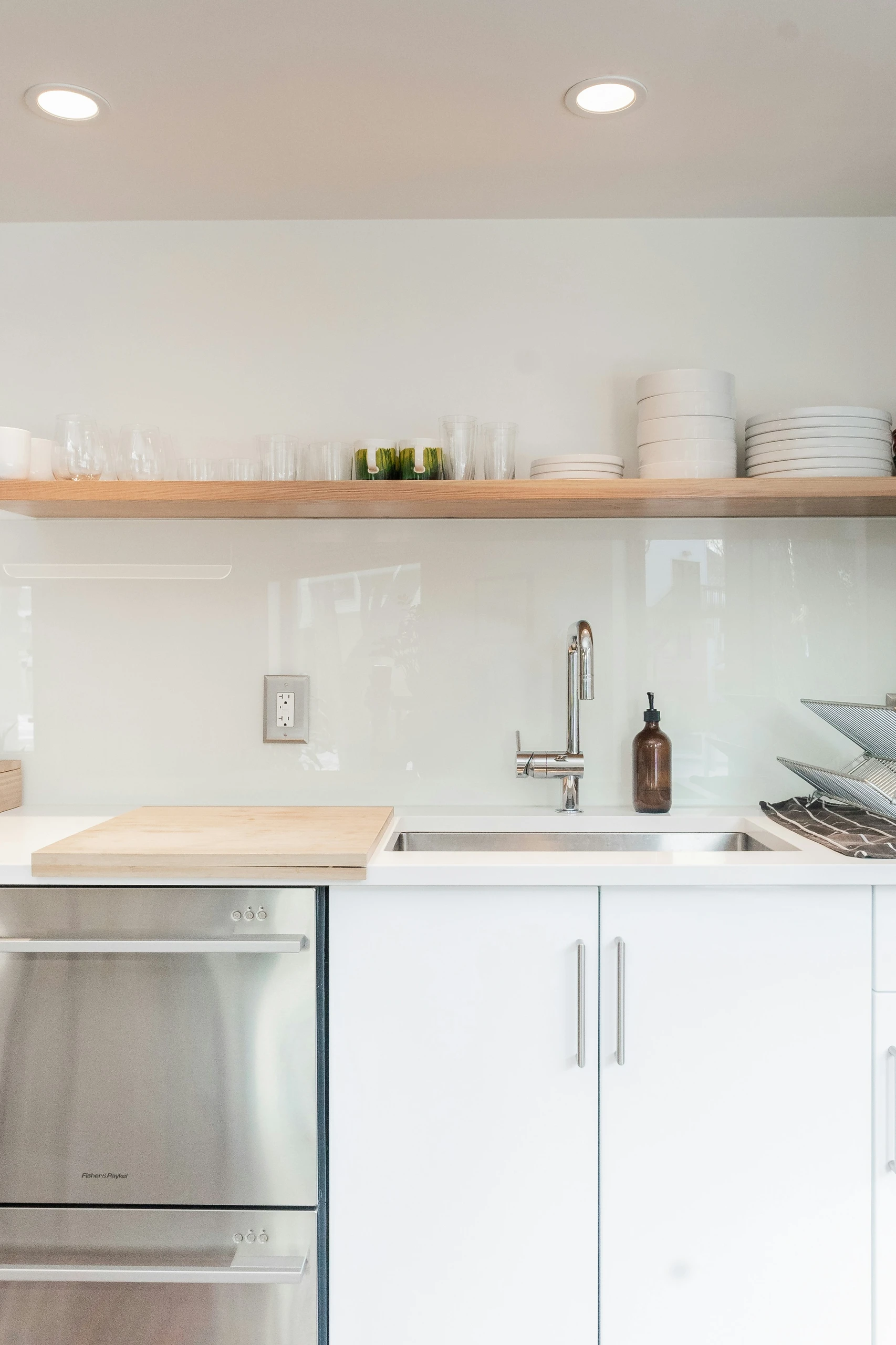 A bright and minimal kitchen counter organization idea featuring a clean white countertop with a stainless steel sink, wooden cutting board, and a brown soap dispenser. Above the counter, a sleek wooden floating shelf is neatly arranged with stacked white plates, bowls, and clear glasses, demonstrating how to install floating shelves over your kitchen counter for storage. The uncluttered setup maximizes space while keeping everyday essentials within easy reach, blending functionality with modern style.