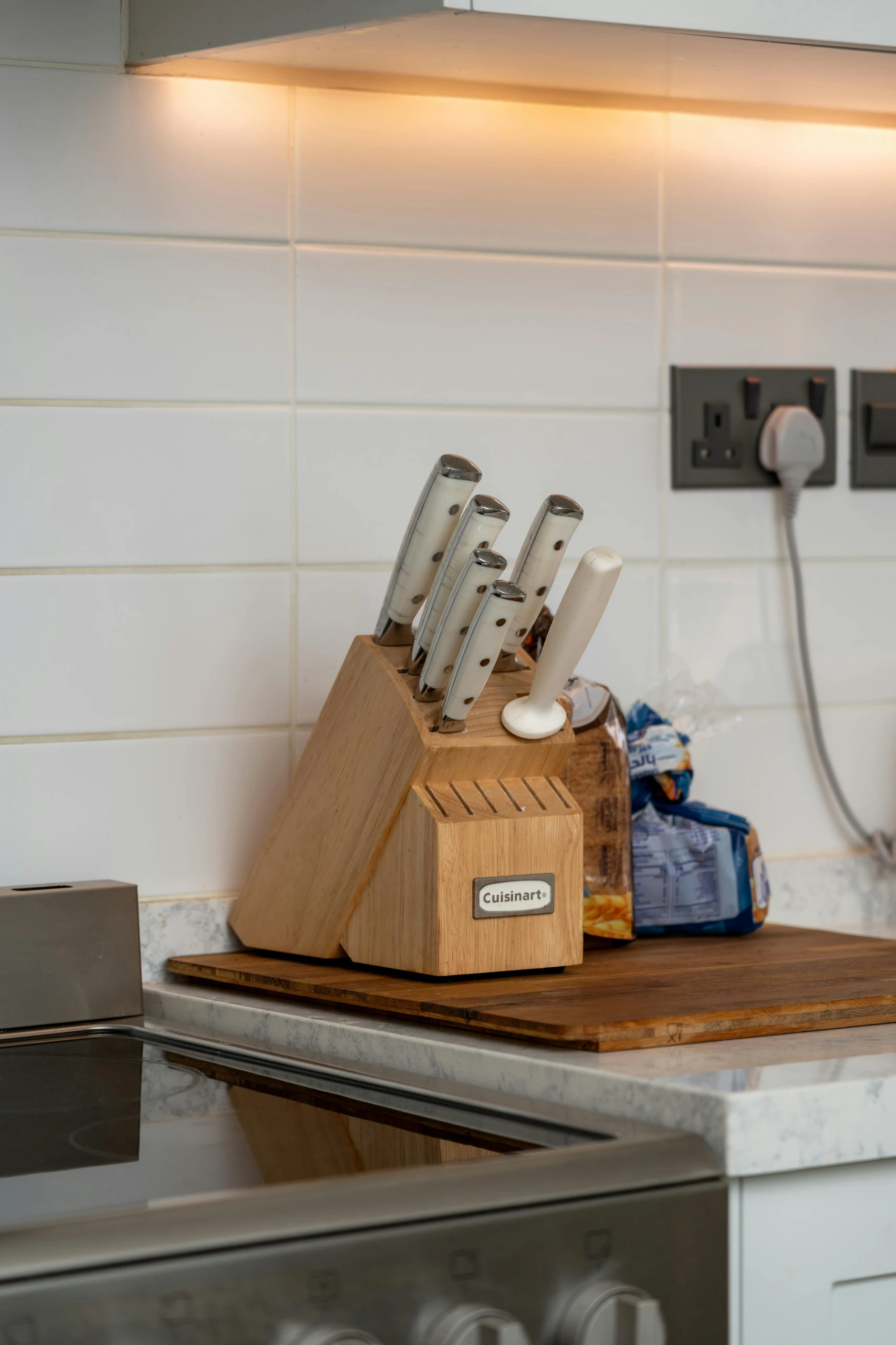 A modern kitchen scene highlighting smart kitchen counter organization with knifes in a knife block. A sleek wooden Cuisinart knife block, filled with stainless steel knives and a sharpening rod, rests neatly on a wooden cutting board beside a clean white tiled backsplash. Warm under-cabinet lighting and minimal countertop clutter create a versatile, functional, and stylish cooking space.