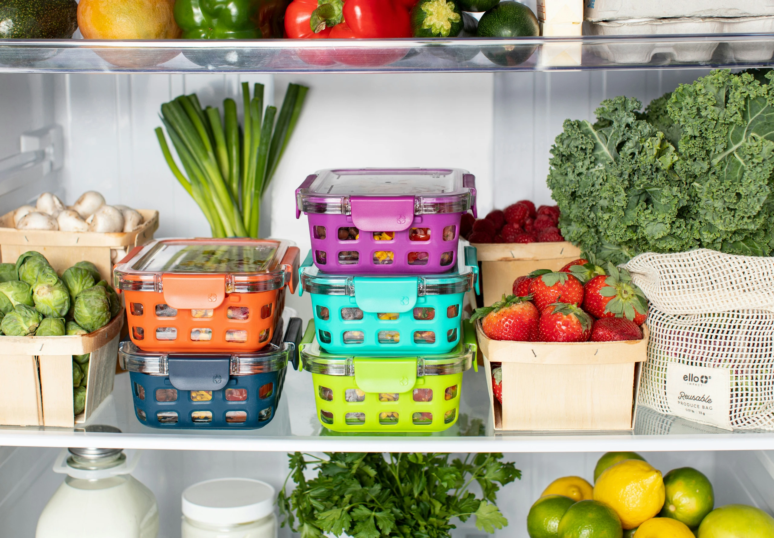 A vibrant and practical kitchen counter organization idea showing how to use stackable boxes to store food neatly. Inside a refrigerator, colorful stackable containers in orange, blue, purple, turquoise, and lime green are filled with fresh ingredients, surrounded by baskets of strawberries, Brussels sprouts, mushrooms, leafy greens, and reusable produce bags. The setup keeps produce visible, organized, and easy to access while maximizing storage space.