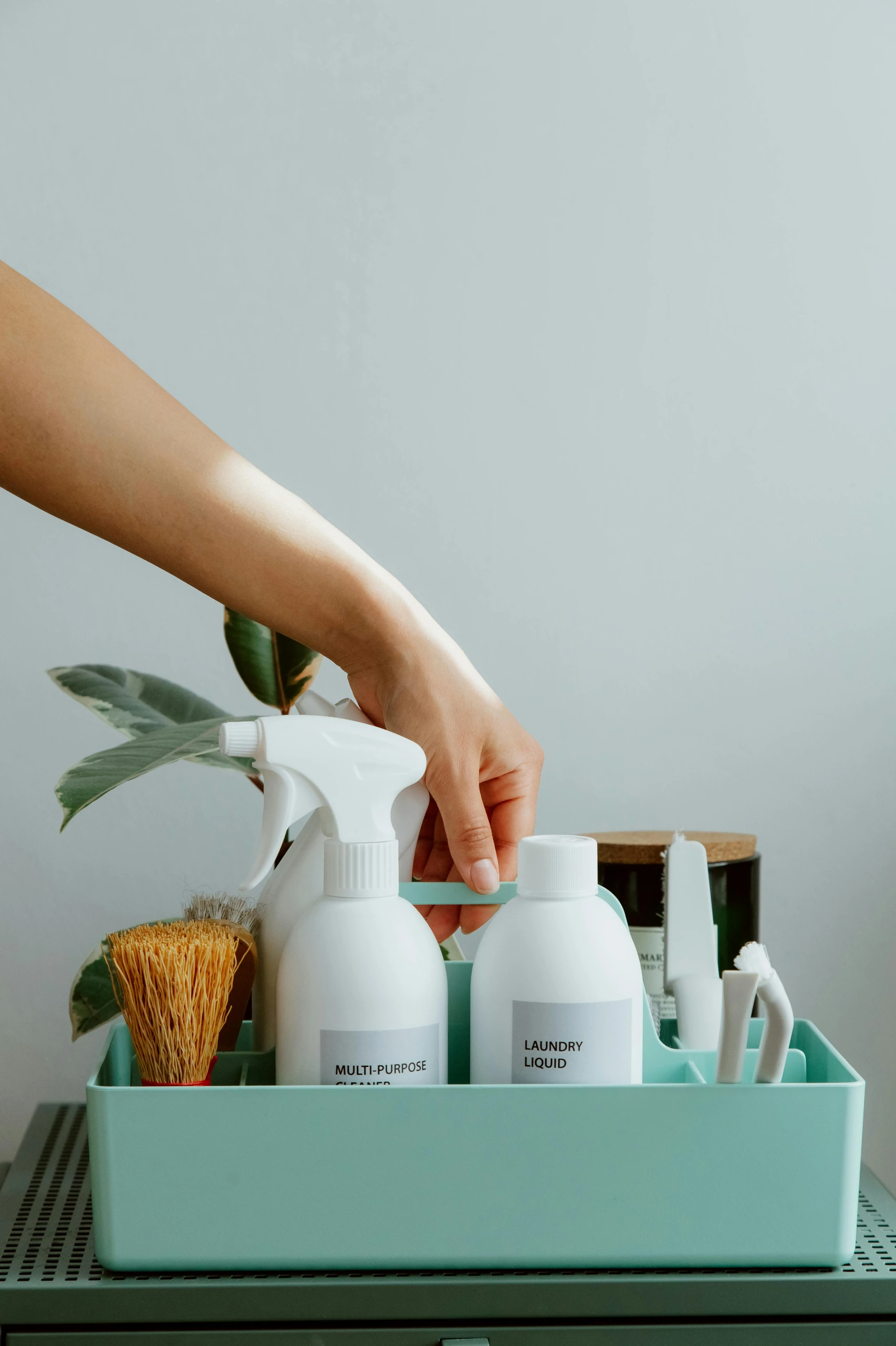Practical kitchen counter organization idea showing how to stow your cleaning tools neatly in a compact caddy. A hand reaches for a mint-green storage container holding a multi-purpose spray bottle, laundry liquid, a scrub brush, and small cleaning accessories. The setup is simple yet efficient, keeping everyday cleaning supplies within easy reach while maintaining a tidy and uncluttered counter space.