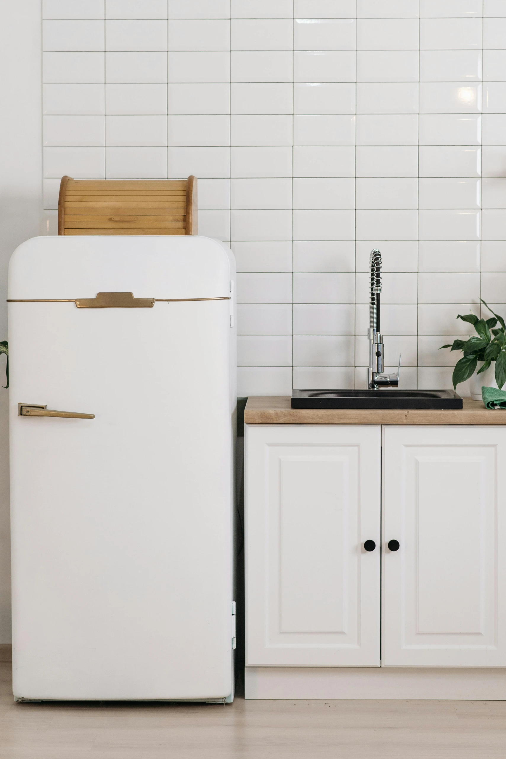 Minimalist kitchen setup showcasing smart kitchen counter organization with the idea to use the top of your fridge for storage. A vintage-style white refrigerator holds a wooden bread box on top, next to a compact sink area with white cabinets, a light wood countertop, a modern chrome faucet, and a small green plant for a fresh touch. The clean white tiled backsplash and uncluttered surfaces create a bright, airy, and functional kitchen space.