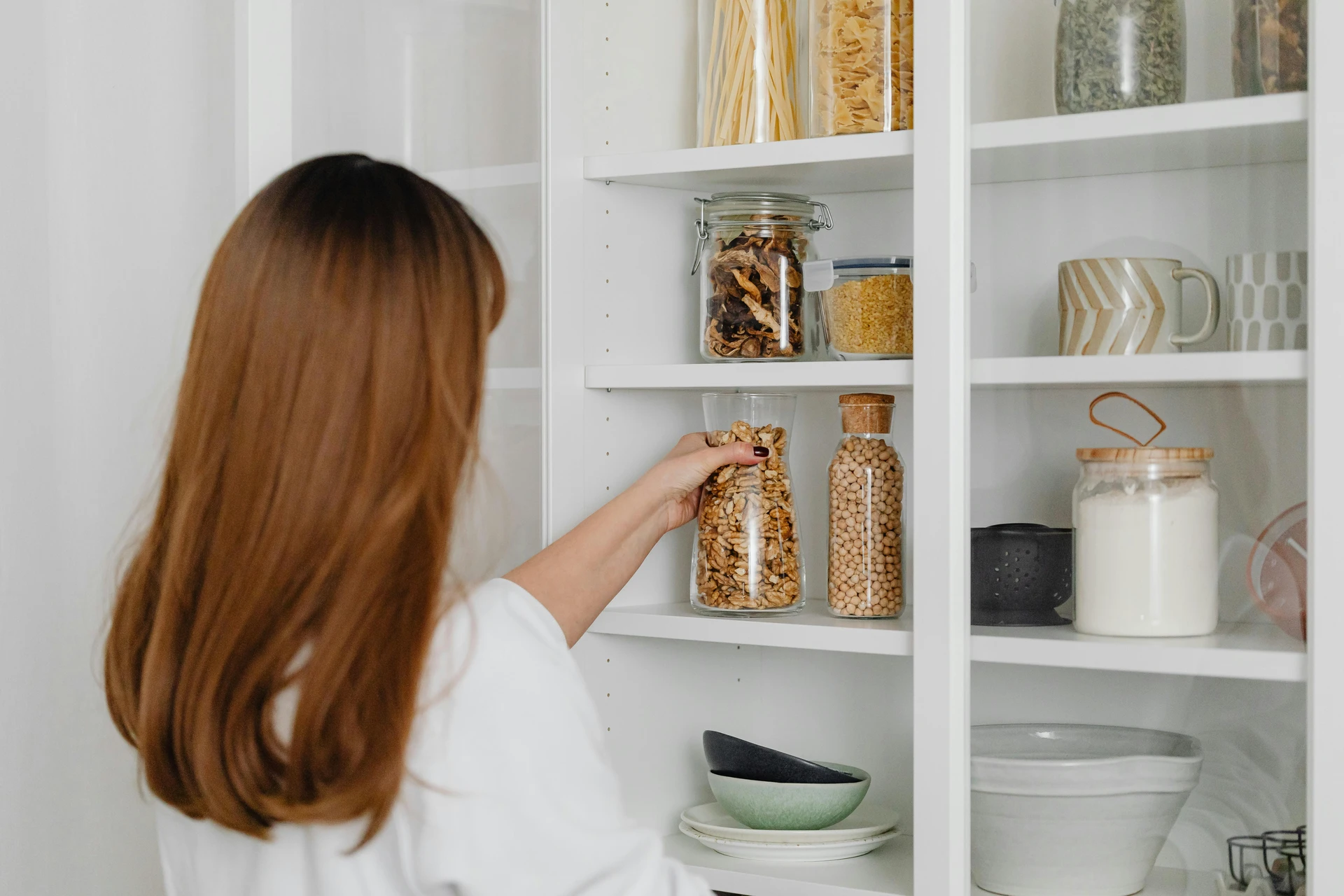 A woman organizing pantry shelves by placing dry goods in stylish clear jars, showcasing a practical and aesthetic way to store pasta, grains, and legumes. This approach is a great example of kitchen organization ideas for small kitchens, where you can use clear jars to save space, keep ingredients visible, and add a decorative touch to open or glass-front cabinets.