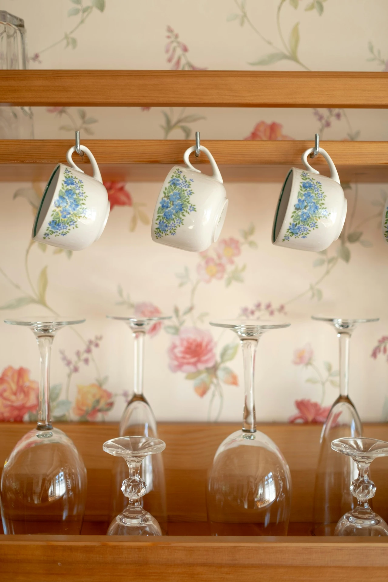 A wooden kitchen rack with floral teacups neatly hang cups on hooks above an arrangement of upside-down wine glasses, set against a charming floral wallpaper. This clever display combines style and function, making it a perfect example of kitchen organization ideas for small kitchens, where vertical space is maximized for both storage and decoration.