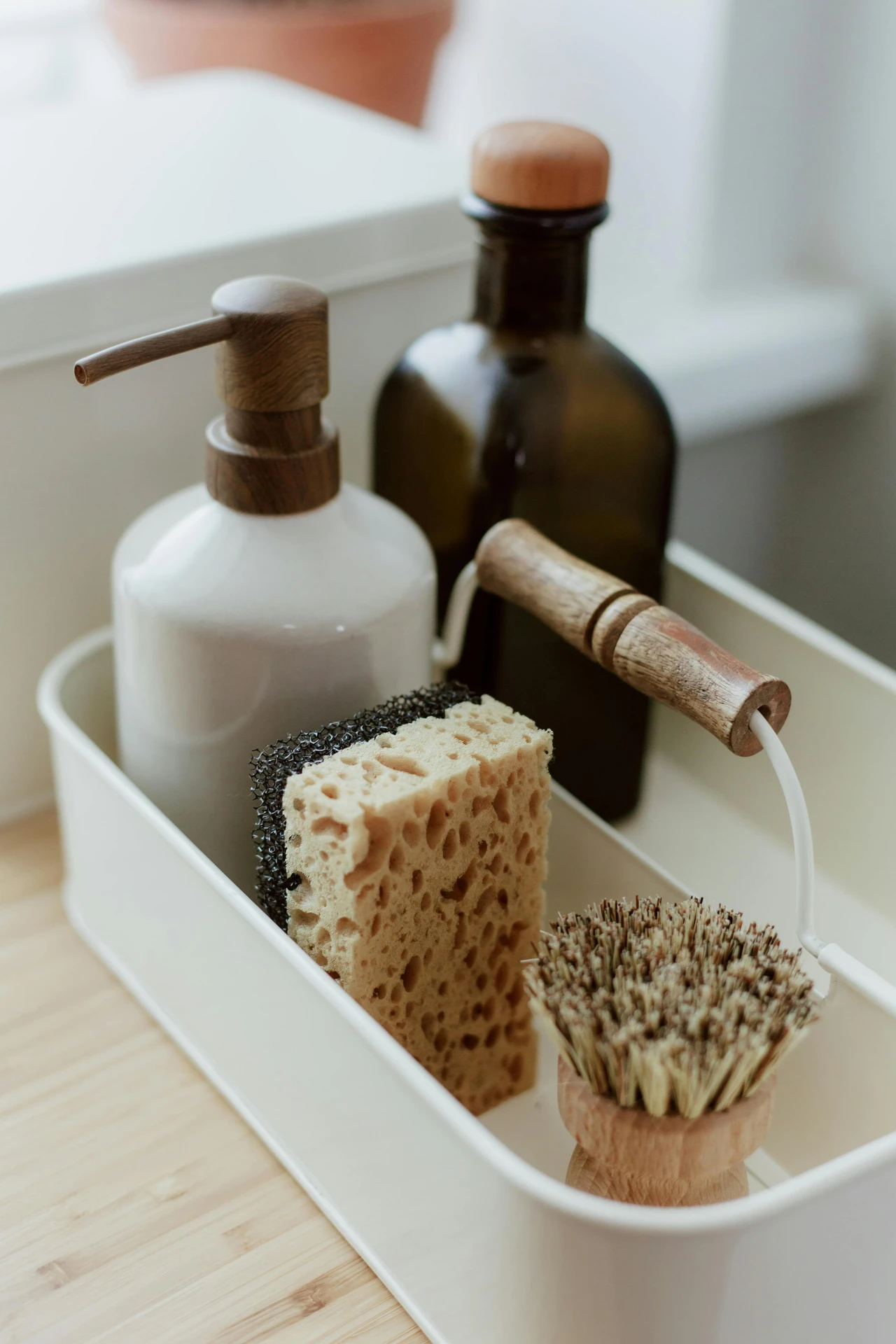 Neatly arranged dishwashing essentials in a compact white caddy, featuring a soap dispenser with a wooden pump, a dark glass bottle with a wooden lid, a sponge, and a natural bristle brush. Perfect for those looking to keep your dishwashing utensils in boxes, this setup offers practical and stylish kitchen organization ideas for small kitchens, ensuring a clutter-free and efficient workspace.