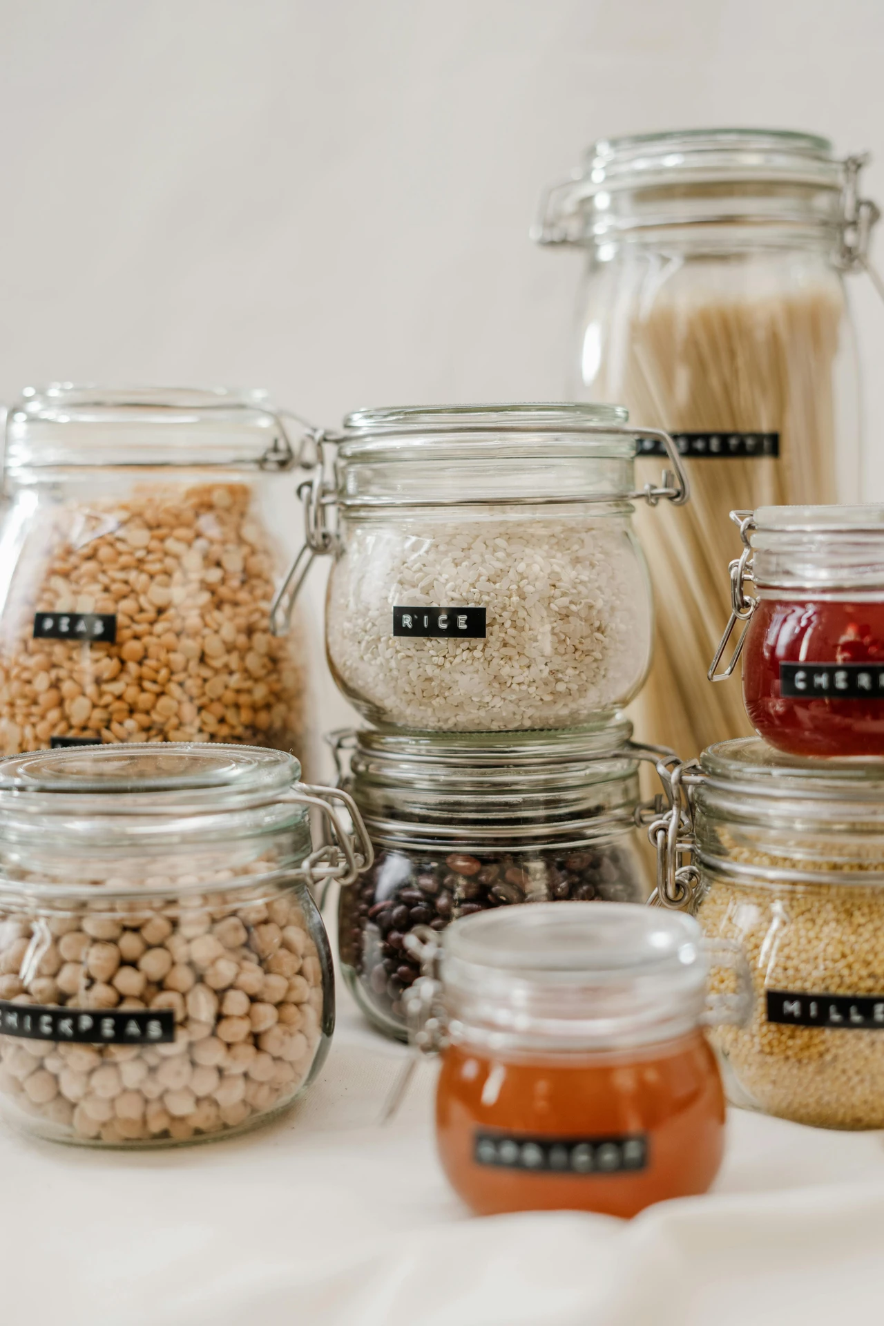 A neatly arranged collection of clear glass jars filled with various pantry staples, each labeled with black and white tags such as “rice,” “chickpeas,” and “millet.” The jars hold pasta, grains, legumes, dried fruit, and honey, showcasing how to label jars and boxes for quick identification. This setup is a stylish and practical example of kitchen organization ideas for small kitchens, helping maximize space, keep ingredients fresh, and maintain a tidy, visually pleasing pantry.