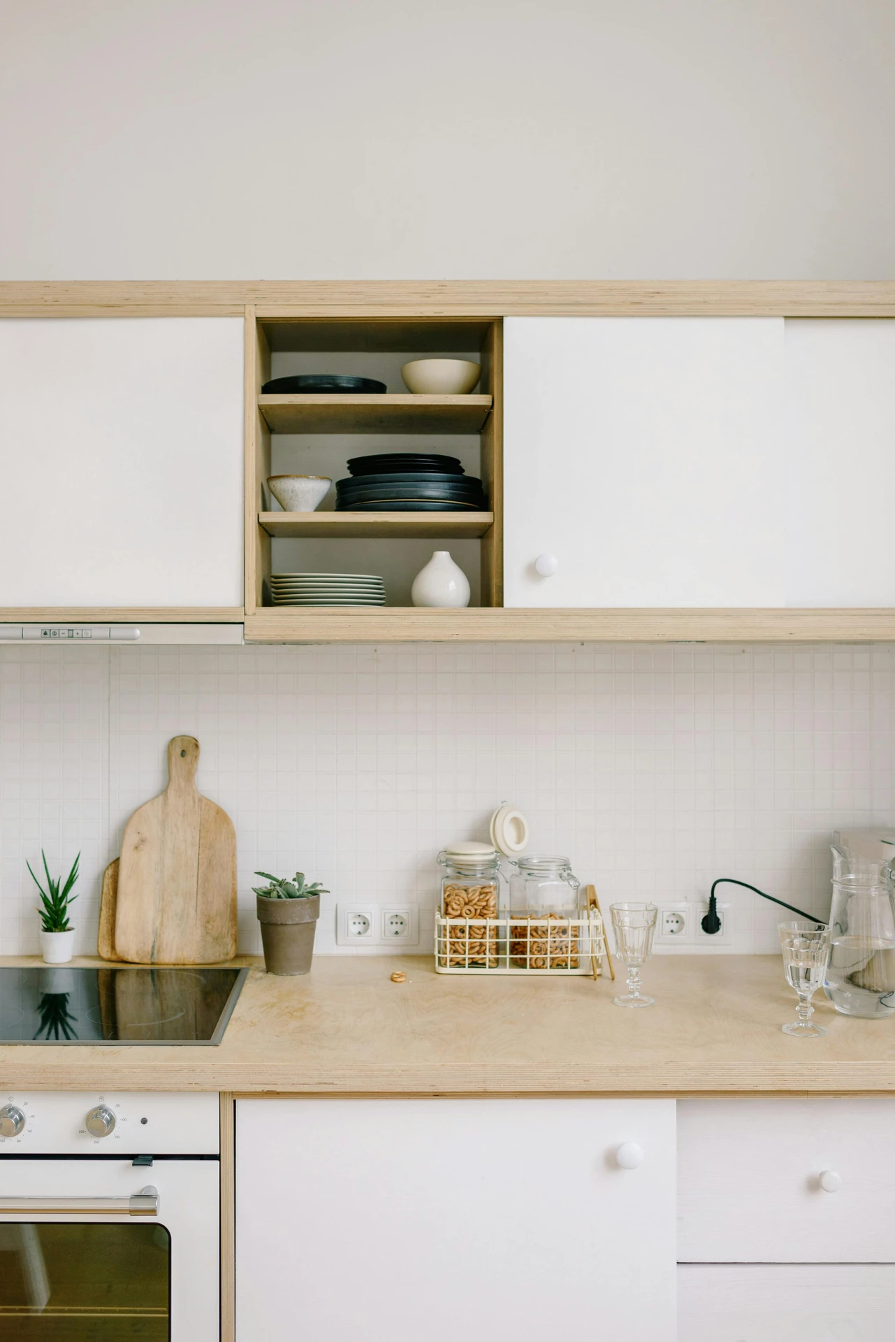 Minimalist kitchen with an organized countertop, featuring glass jars filled with snacks, a wooden cutting board, and small potted plants for a touch of greenery. Open shelving above displays neatly stacked plates, bowls, and decorative ceramics. This clean, functional setup is a great example of kitchen organization ideas for small kitchens, maximizing space while keeping essentials accessible and maintaining a visually appealing, clutter-free look.