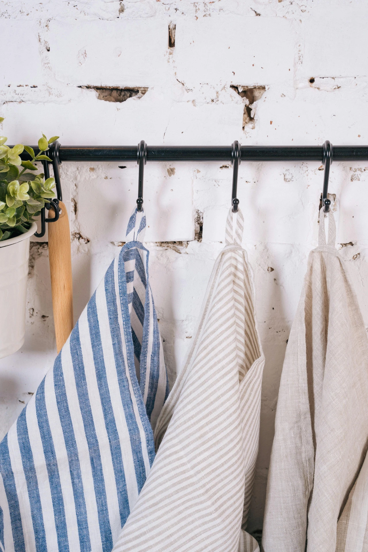A black metal rod mounted against a rustic white brick wall, featuring three striped tea towels in blue, beige, and neutral tones neatly hanging on individual hooks, with a potted plant and wooden kitchen utensil to the side. A practical way to hang tea towels on hooks, combining easy access with style. Perfect for kitchen organization ideas for small kitchens, adding charm while saving counter and drawer space.