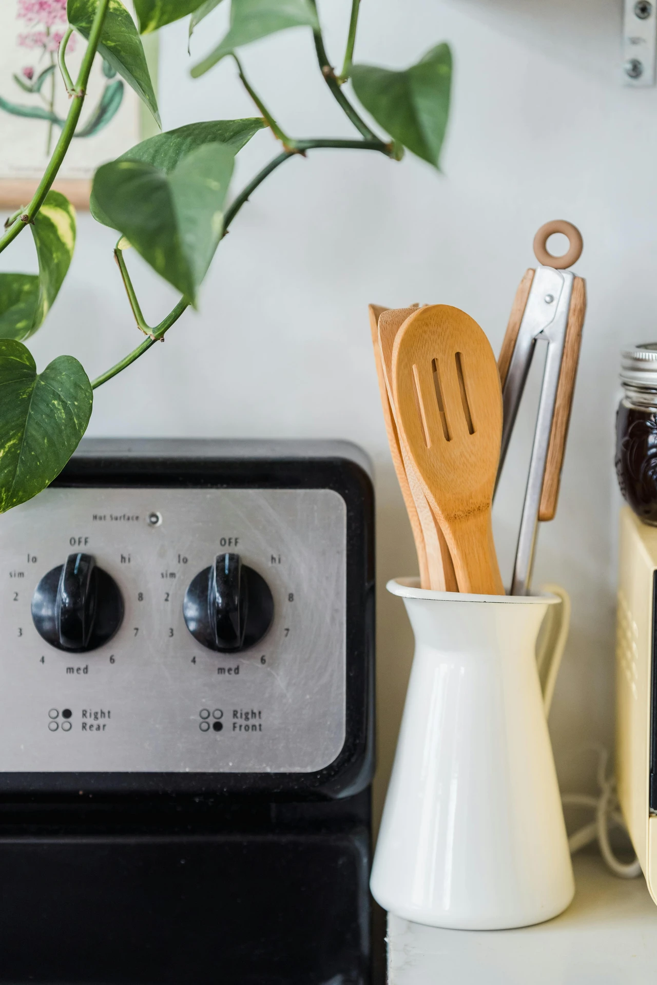 A white ceramic jar holding wooden spoons and metal tongs placed beside a stovetop, demonstrating how to use jars for your utensils as part of smart kitchen organization ideas for small kitchens. This versatile storage solution keeps cooking tools within easy reach while adding a clean, minimalist touch to the kitchen décor.