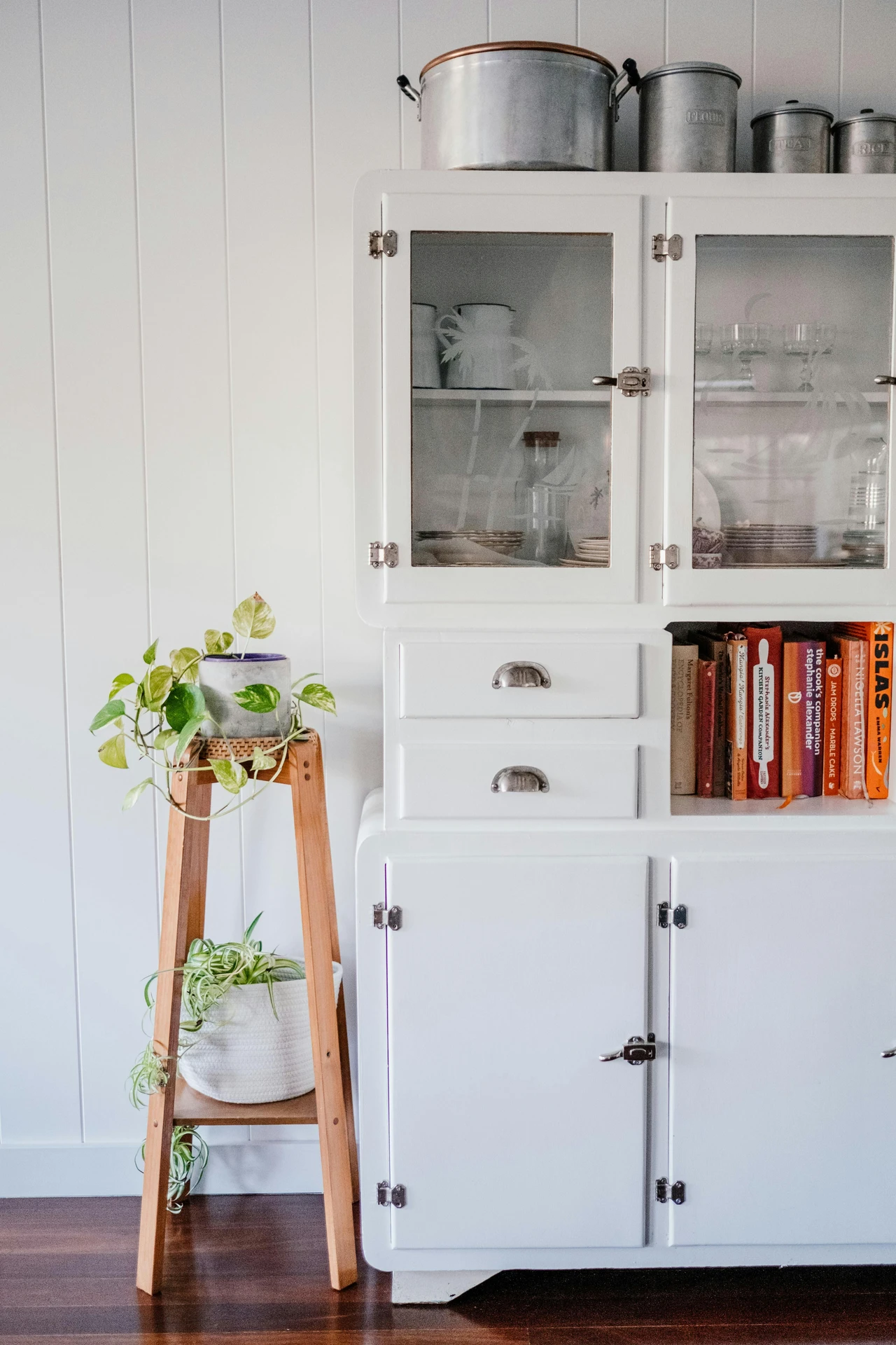 A white vintage-style kitchen cabinet with glass doors neatly displaying dishes and glassware, alongside a small open shelf for cookbooks. On the left, a wooden plant stand holds two potted plants, adding greenery and charm. This setup is a creative example of vertical storage, combining aesthetics with function, and a smart choice for kitchen organization ideas for small kitchens where maximizing space while keeping items accessible is essential.