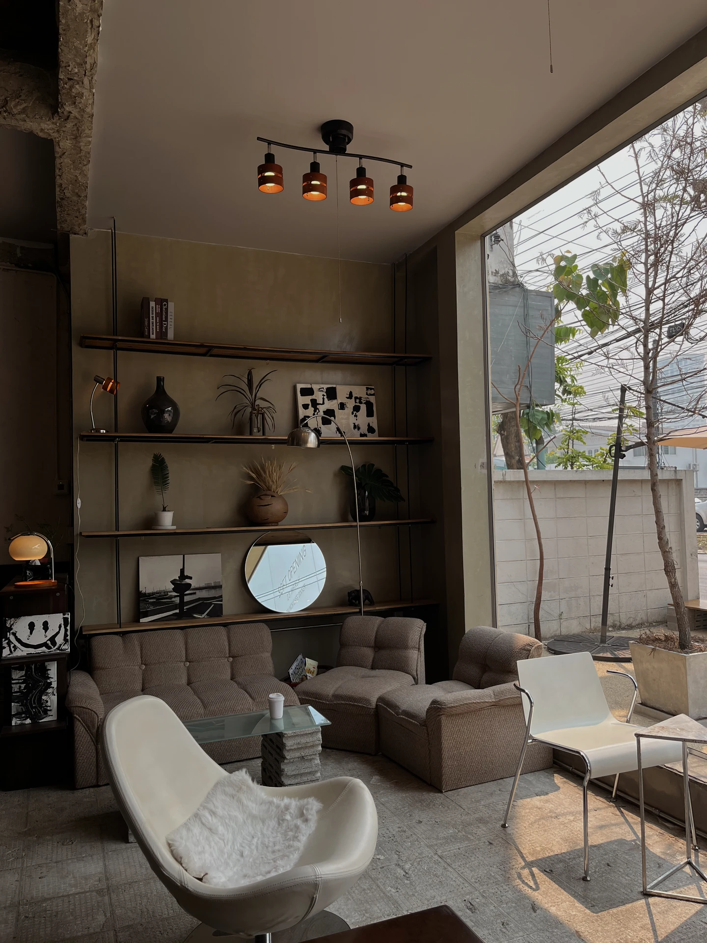 Industrial-style living room with floor-to-ceiling windows, featuring a low modular brown sofa, a white modern accent chair with a fur throw, and a sleek glass coffee table. Open shelving with eclectic decor and warm ceiling lights add character. The arrangement embraces asymmetry modern design, mixing different seating styles and heights for a relaxed yet stylish look. This setup is a creative example of living room layout ideas, showing how asymmetry can make a space feel dynamic and personalized.