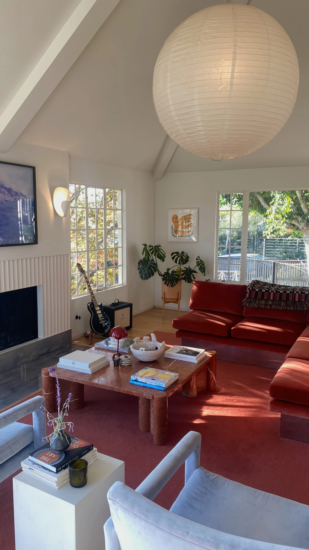Bright and eclectic living room with vaulted ceilings, a bold red sectional sofa, soft gray armchairs, and a large stone coffee table at the center, styled with books and decor. The seating forms a conversation circle, encouraging interaction while keeping the fireplace and TV in view. Large windows bring in natural light and greenery, adding warmth to the space. A versatile example of living room layout ideas, showing how a circle of seating creates both intimacy and flexibility for gatherings.