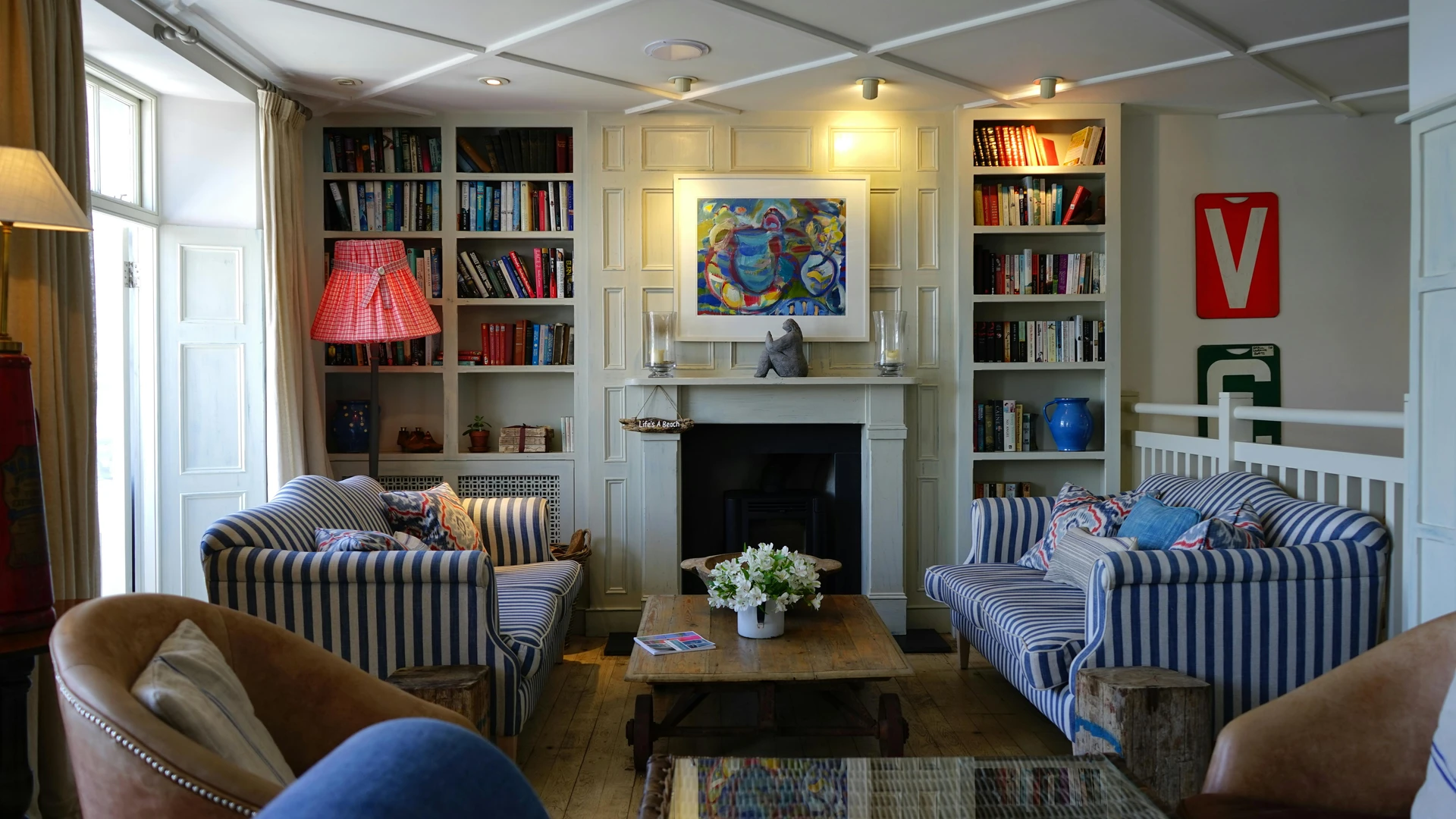 Cozy living room layout ideas with face-to-face sofas. Two striped blue-and-white sofas are arranged opposite each other with a rustic wooden coffee table in between, creating an inviting conversation area. Built-in bookshelves frame a fireplace with artwork above, while neutral curtains, accent chairs, and a mix of colorful pillows add charm and versatility to the space.