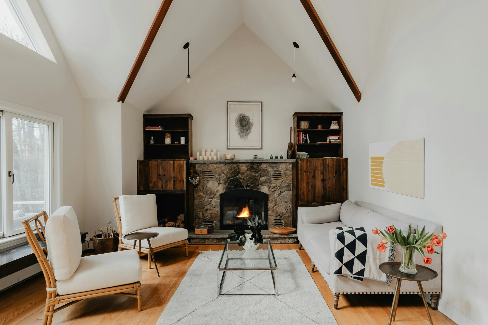 Warm and inviting living room with vaulted ceilings and exposed beams, featuring a stone fireplace framed by wooden cabinets and styled with candles and artwork. A light gray sofa and two rattan armchairs create a cozy fireplace arrangement around a glass coffee table and soft rug. This versatile example of living room layout ideas shows how seating can be oriented around a focal point, making the space perfect for both everyday relaxation and social gatherings.