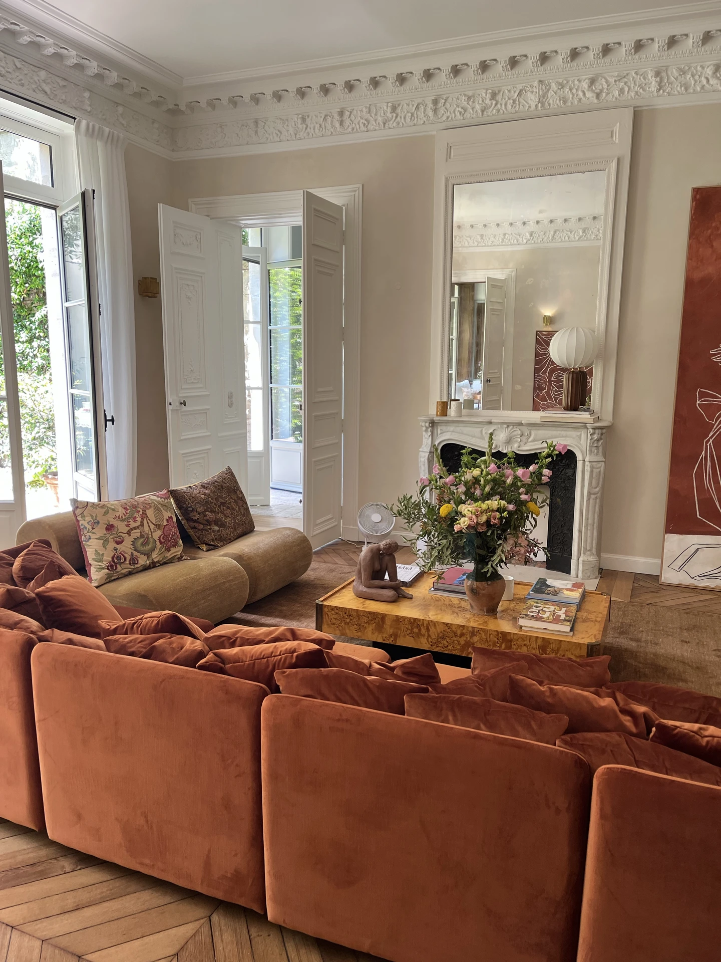 Elegant living room with ornate crown molding, tall French doors opening to a garden, and a striking mix of seating including a burnt orange velvet sofa and a rounded beige armchair. The arrangement forms a floating island around a wooden coffee table decorated with flowers and books, leaving space around the furniture for easy flow. This setup is a great example of versatile living room layout ideas, showing how a floating island arrangement can work in both classic and modern interiors, creating a cozy yet open conversation zone.