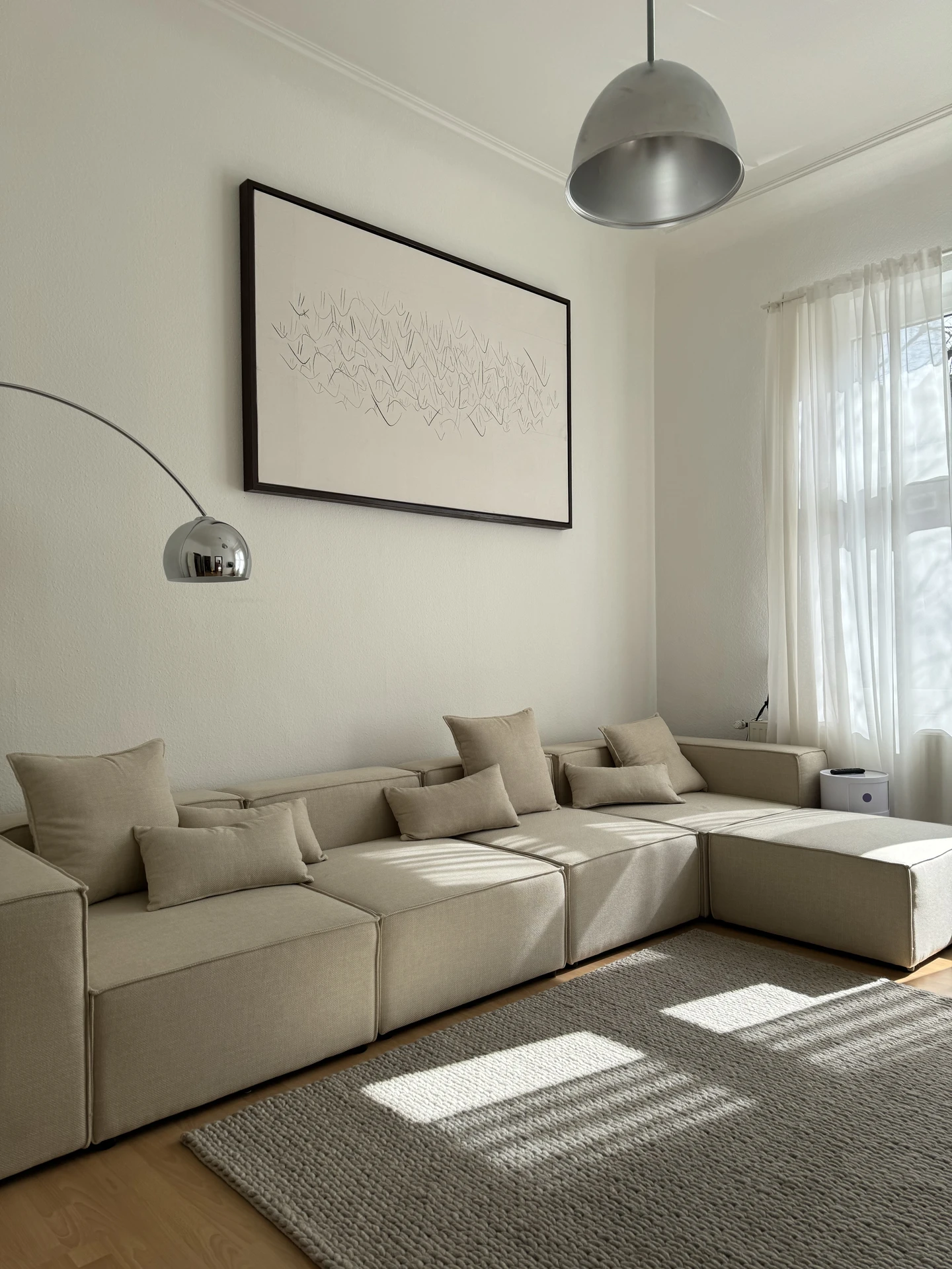 Bright and minimalist living room with neutral tones, featuring a modern l-sectional sofa in beige with multiple cushions, a textured area rug, sheer white curtains, and metallic pendant lamps. The clean layout demonstrates cozy simplicity but also works as inspiration for versatile living room layout ideas, as the modular sectional can be rearranged for different seating styles and room sizes.