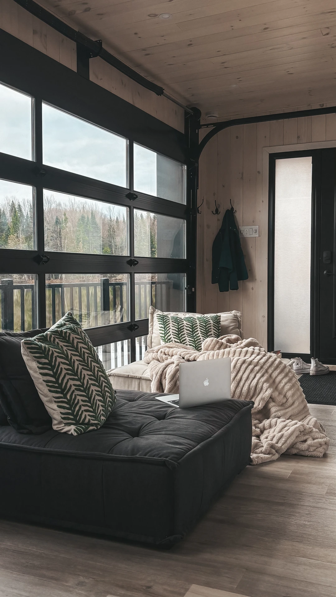 Cozy modern living space with large industrial-style windows, light wood walls, and a pair of cushioned loveseat pair seats topped with patterned pillows and a soft throw blanket. A laptop rests casually on one seat, making the setup perfect for both work and relaxation. This arrangement is a versatile example of living room layout ideas, showing how a simple loveseat duo can create an intimate, functional seating area in small or open-plan spaces.
