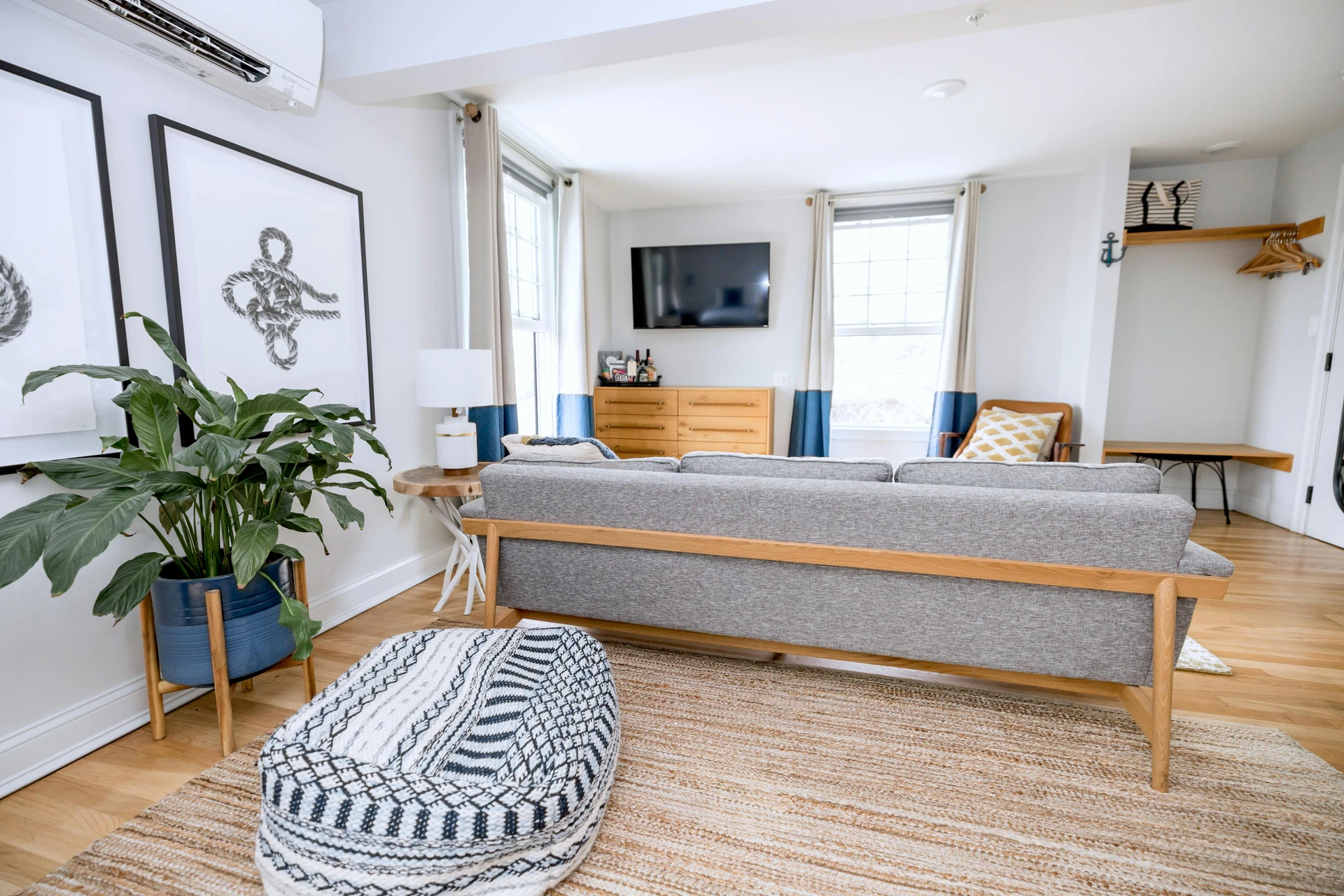 Bright and airy living space with a mid-century style gray sofa, patterned pouf, woven rug, and indoor plant adding a touch of greenery. A wall-mounted TV sits above a wooden dresser, while accent chairs and layered textiles create a casual, relaxed vibe. This cozy low-lounge den is a versatile example of living room layout ideas, showing how simple furniture and soft seating arrangements can make a space feel both functional and inviting for everyday comfort.