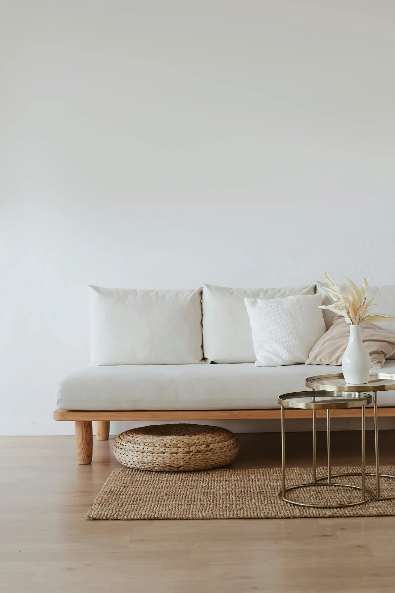 Minimalist living space with a light wood floor, a simple white sofa with soft neutral pillows, and a woven pouf tucked underneath. In front, sleek metallic nesting tables instead of one coffee table hold a white vase with dried foliage, creating a flexible and airy setup. This is a versatile example of living room layout ideas, showing how multi-use furniture like nesting tables can maximize space while keeping the design elegant and functional.