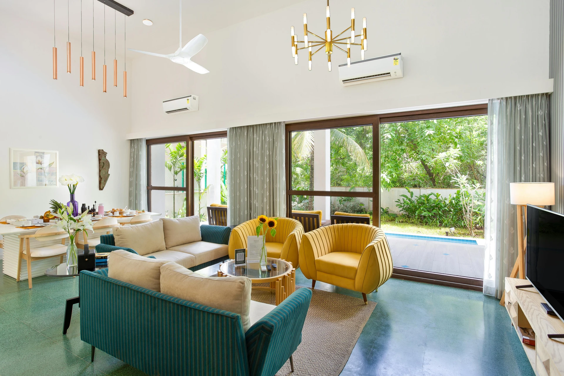 Bright and modern living room with floor-to-ceiling windows opening to a lush garden, featuring two matching teal sofas and a pair of bold yellow armchairs arranged around a glass coffee table. The setup uses symmetry classic design principles, balancing color and form to create a harmonious, welcoming space. This versatile example of living room layout ideas shows how symmetry can be applied in both contemporary and traditional settings, making the room feel structured yet inviting.