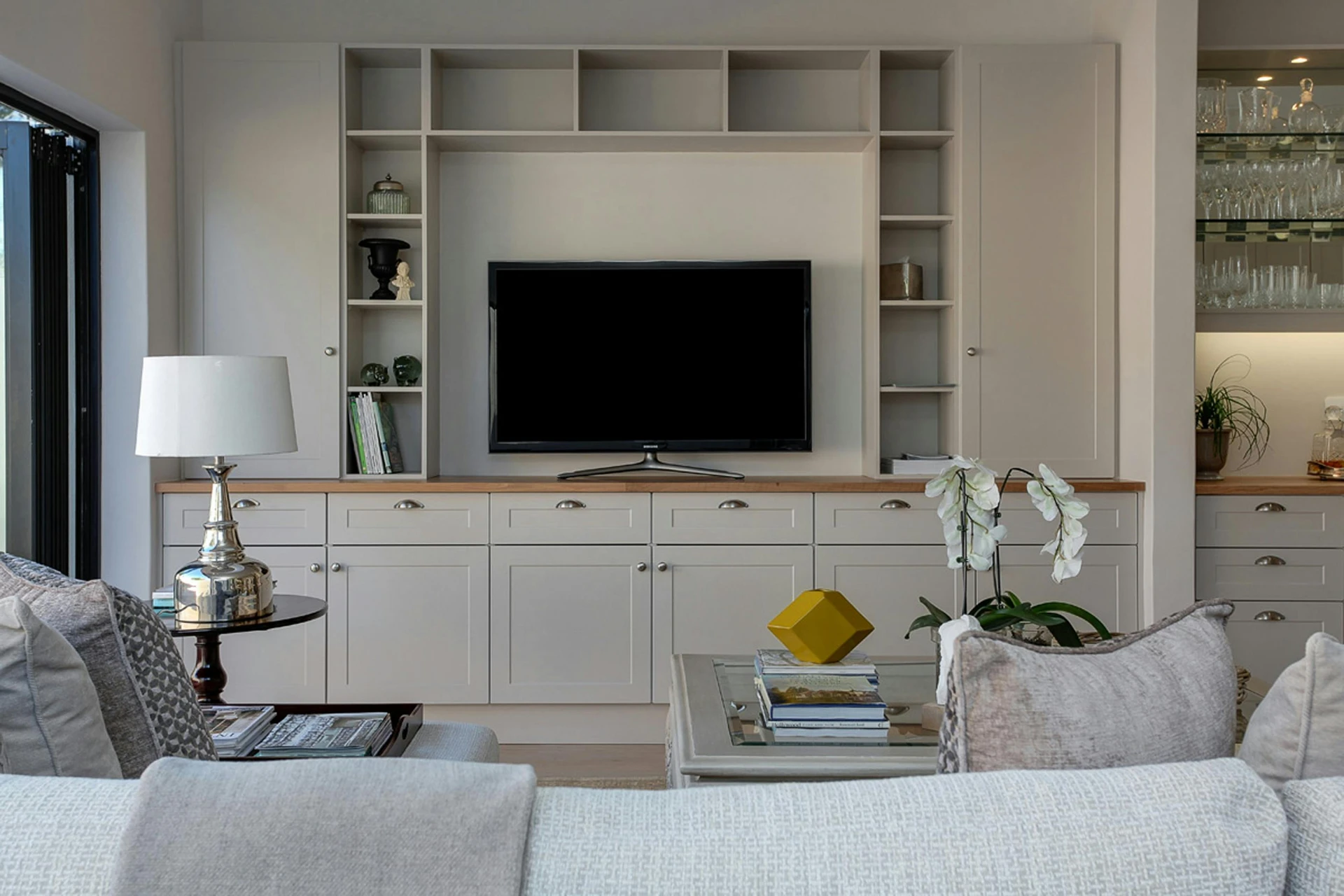 Modern living room with built-in shelving and cabinetry framing a flat-screen TV, complemented by neutral-toned sofas, a glass coffee table with books and decor, and a side table with a chrome lamp. This setup highlights a tv-corner layout, where seating is oriented for comfortable viewing without overwhelming the room's design. A versatile example of living room layout ideas, it balances function and style, making the TV a focal point while keeping the space elegant and inviting.