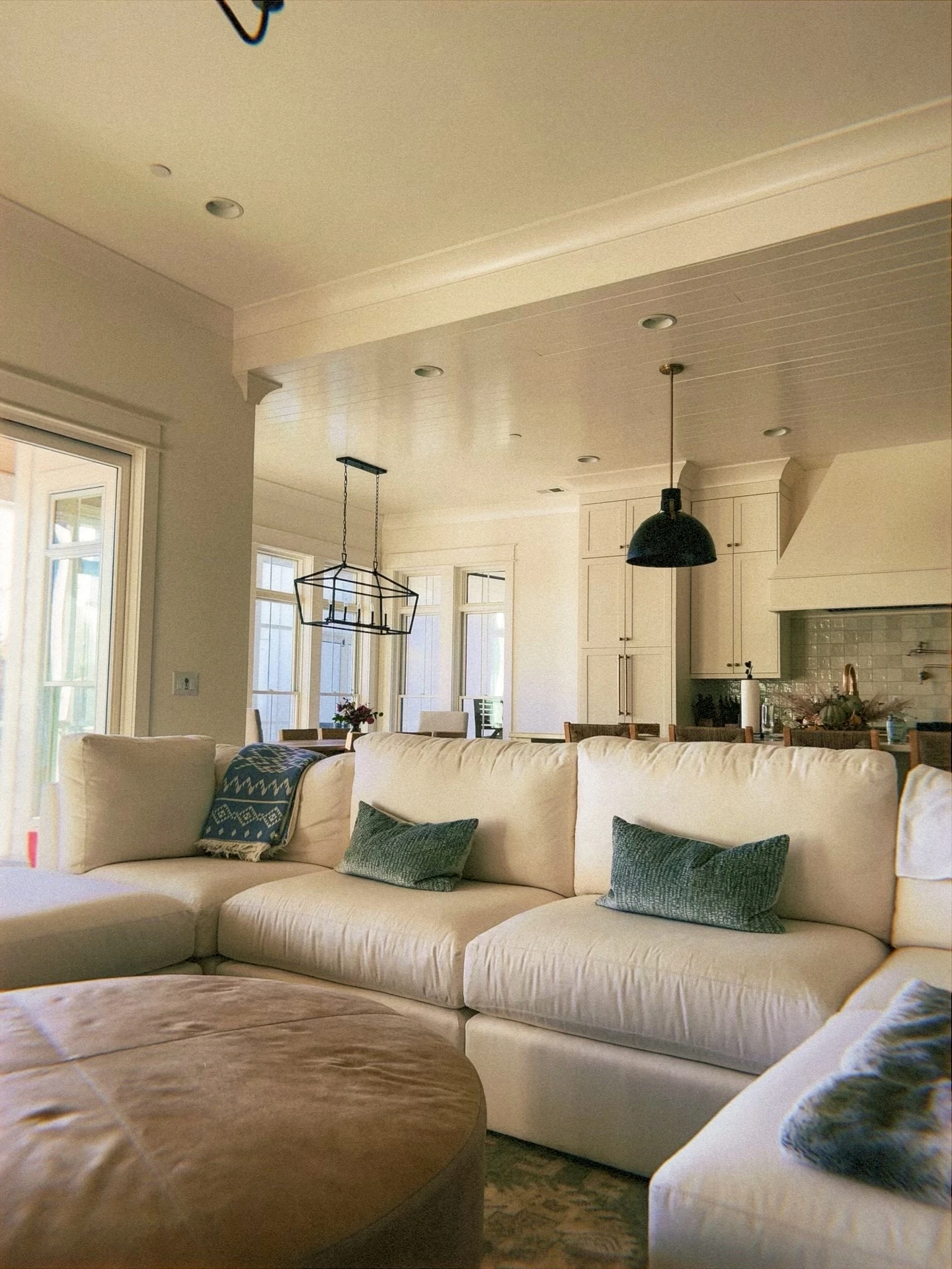 Cozy open-plan living room with a spacious u-shape sofa in soft beige, styled with green and blue accent pillows, a patterned throw, and a large round ottoman serving as a central coffee table. Natural light flows in through tall windows, while the space seamlessly connects to the kitchen and dining area. This setup highlights versatile living room layout ideas, where a u-shaped seating arrangement encourages conversation, relaxation, and an easy flow between lounging and entertaining.