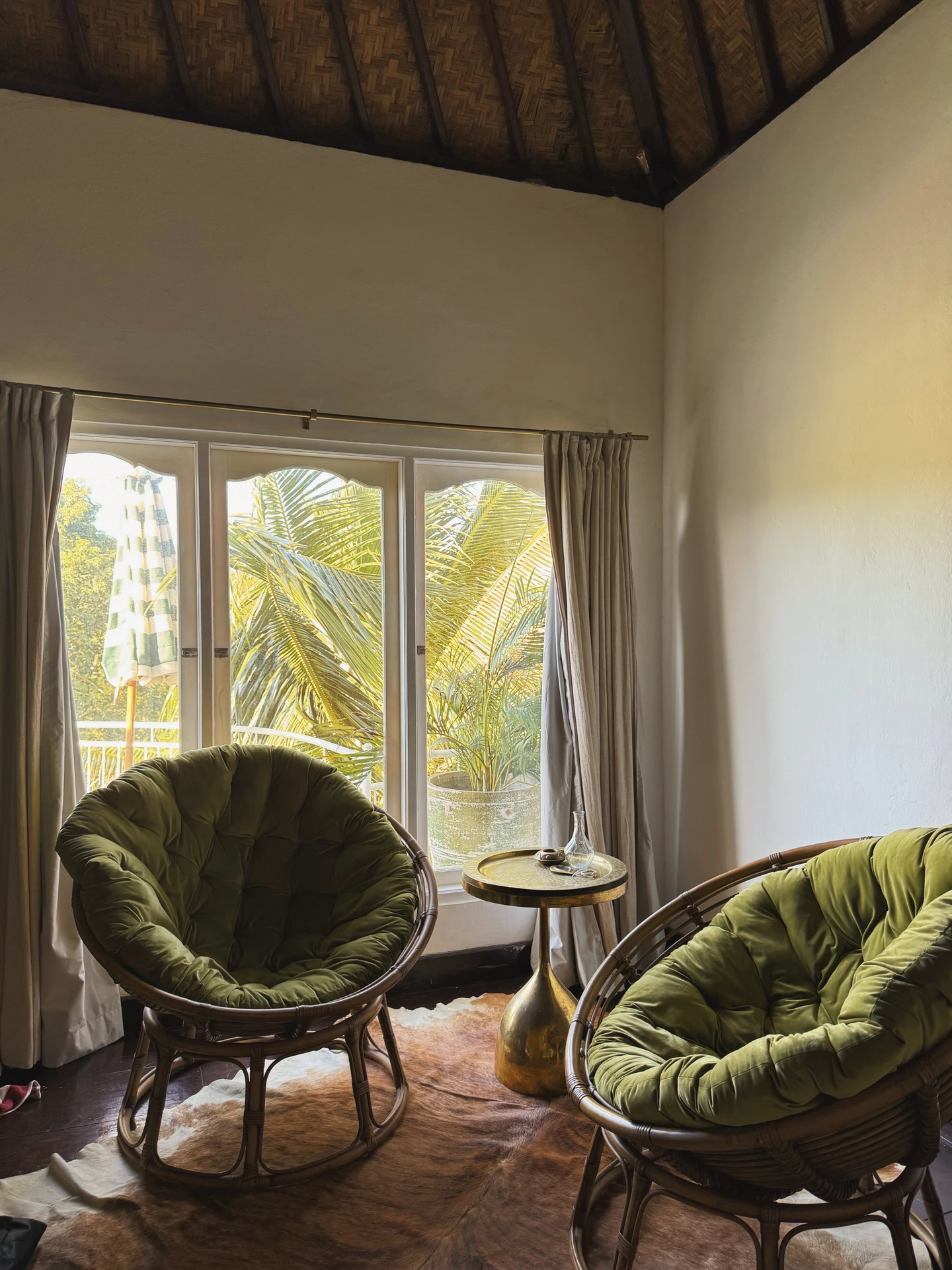 Cozy tropical corner with two rattan papasan chairs featuring deep green cushions, a golden side table, and large glass doors opening to palm trees outside. The setup creates a relaxing window nook that brings nature indoors while offering a perfect spot for reading, coffee, or conversation. This is a versatile example of living room layout ideas, showing how even a small corner by the window can be transformed into a functional and inviting seating area.