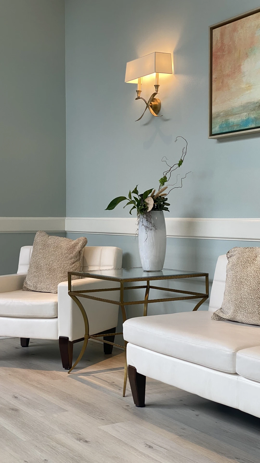 Elegant seating arrangement after a living room makeover, featuring two white armchairs with textured pillows, a gold-framed glass side table, and a tall vase with greenery. The soft blue accent wall is enhanced by a glowing wall sconce and abstract wall art, creating a calm yet refined backdrop that ties the whole room together.