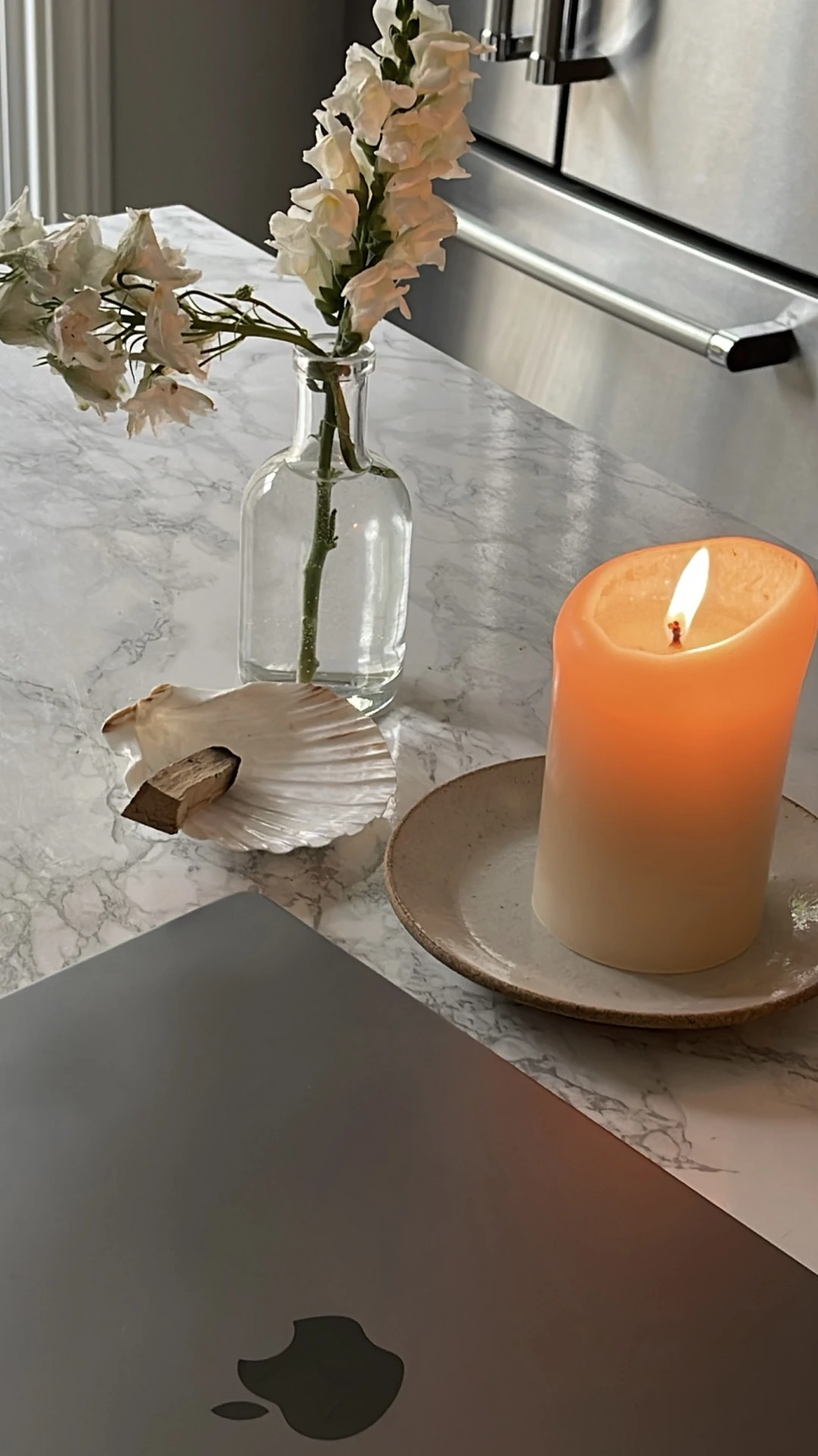 Cozy living room makeover detail with a marble-look surface styled with a glowing pillar candle on a ceramic plate, a delicate glass vase holding white blooms, and a seashell dish for natural accents. The soft flame adds warmth and serenity, showing how to decorate your space with candles for an inviting and calming atmosphere.