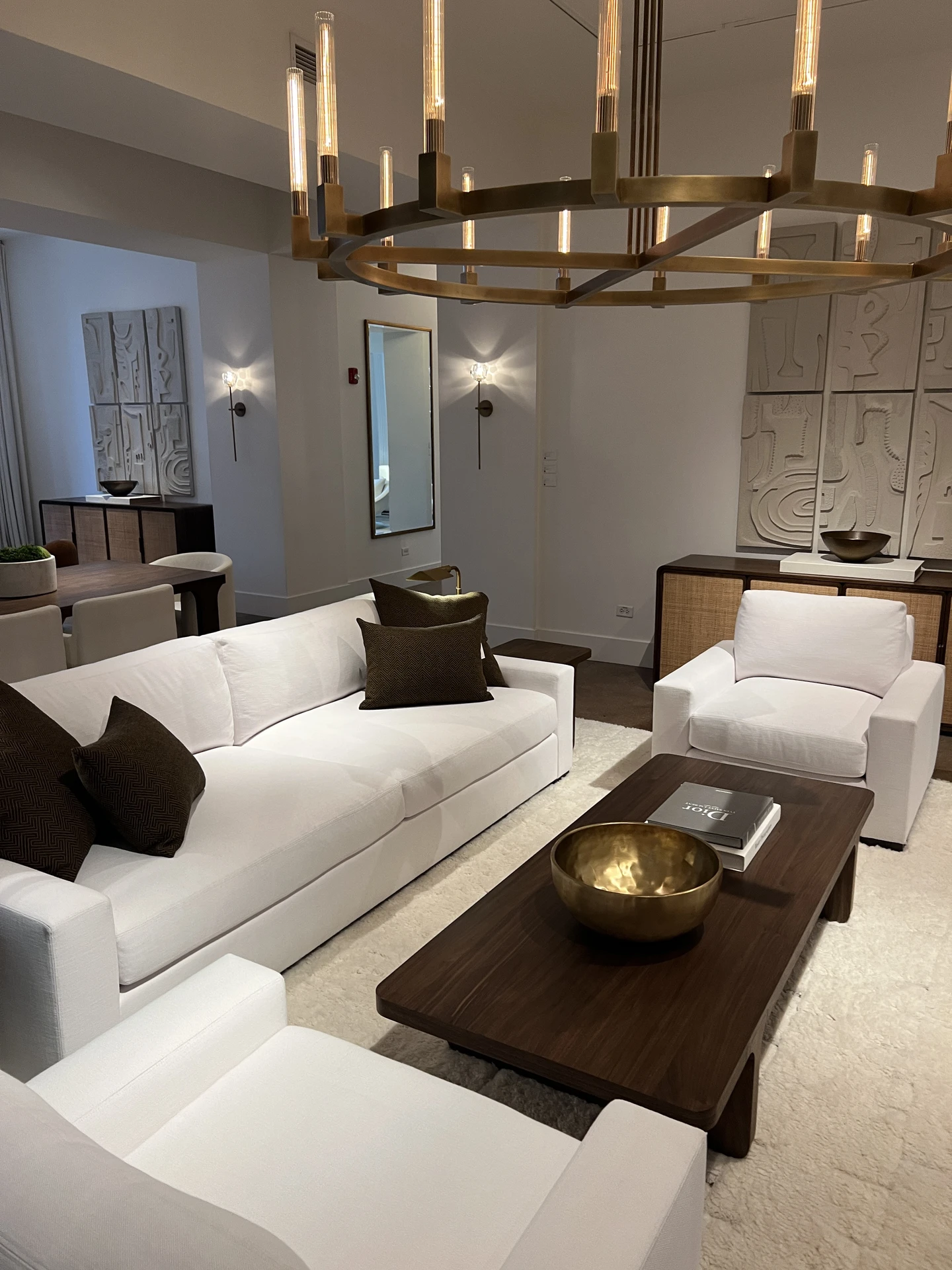 Modern living room makeover featuring crisp white sofas and armchairs accented with dark brown pillows, arranged around a sleek wooden coffee table with a gold bowl and books. A large chandelier and wall art add sophistication, while the clean surfaces and neutral palette show how to declutter your space for a calm, inviting atmosphere. The design highlights balance, order, and understated luxury.
