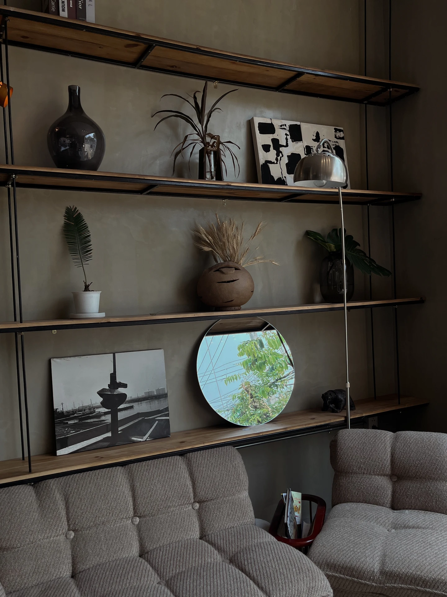 A stylish living room makeover with tufted beige seating and a feature wall showcasing wood-and-metal floating shelves on the wall. The shelves are styled with ceramics, plants, abstract artwork, and a round mirror that reflects greenery from outside, adding depth and character. The layered display creates a warm, curated look that blends modern design with natural touches.
