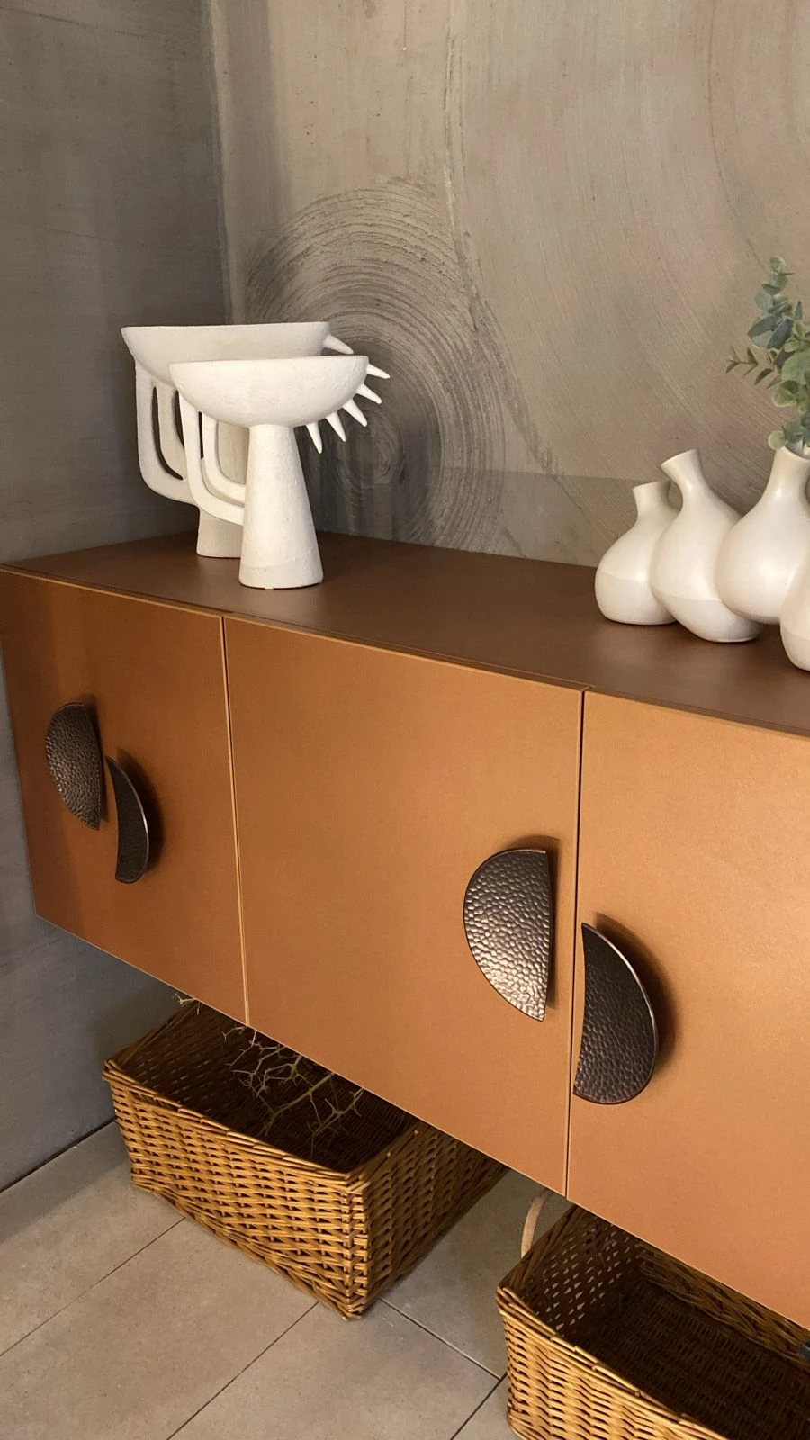 A modern cabinet featured in a living room makeover, showing how to replace old handles with new ones for a stylish update. The sleek bronze cabinet doors are accented with textured crescent-shaped handles, paired with decorative white ceramic vases and sculptural pieces on top. Wicker baskets beneath add both warmth and practical storage, blending functionality with artistic design.