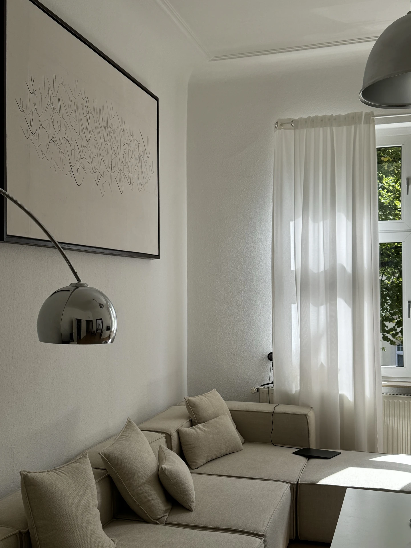 Minimalist living room makeover with a beige modular sofa, neutral cushions, and a sleek chrome floor lamp. Soft white fabric adds elegance as you hang curtains that filter daylight beautifully, creating a calm and airy space. A large abstract artwork on the wall ties the look together with understated sophistication.