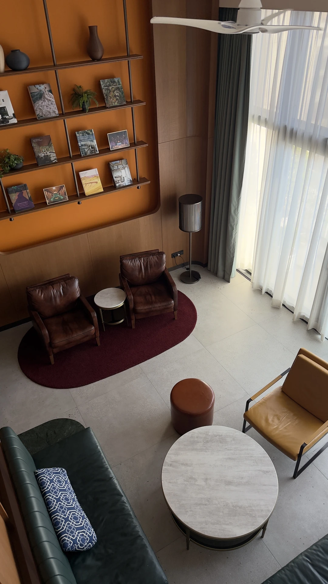 Contemporary loft-style living room makeover with eclectic seating-two brown leather chairs on a burgundy rug, a mustard armchair, a green bench, and a round coffee table at the center. Wall-mounted shelving with books and decor adds character, while tall curtains frame the space with soft light. The arrangement shows how to move furniture around to create flexible conversation zones and highlight unique design elements.