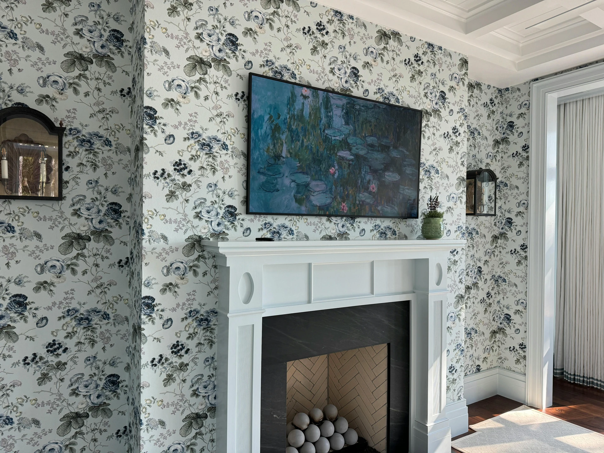 Elegant living room makeover with a white fireplace framed by bold floral new wallpapers in shades of blue, green, and grey. A flat-screen TV doubles as wall art above the mantel, while small decorative touches like a potted plant and vintage wall sconces enhance the cozy yet stylish feel. The patterned walls bring depth and personality, turning the fireplace wall into a striking focal point.