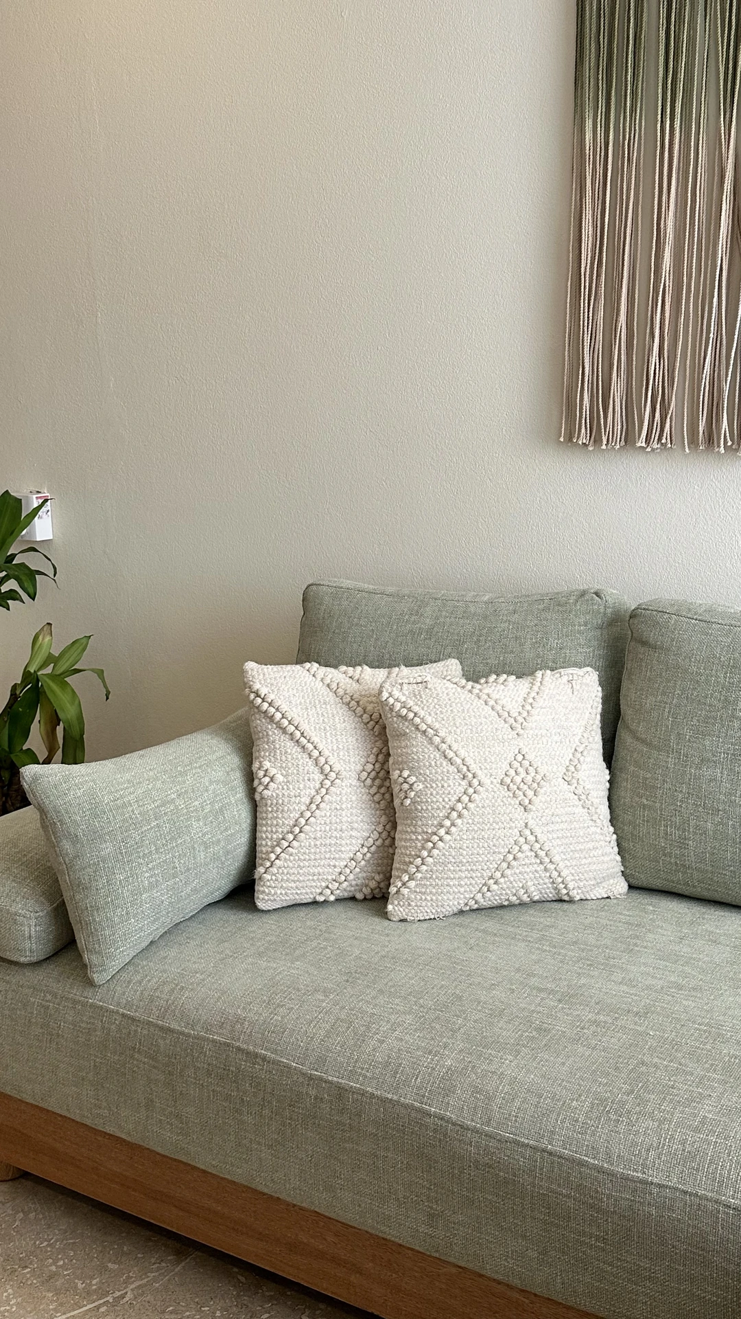 Subtle living room makeover with a sage green sofa featuring two textured cream cushions in a boho pattern. The setup shows how simply you can refresh a space when you replace pillowcases or pillows, instantly adding depth and softness. A touch of greenery and wall-hung macramé complete the calm, natural vibe.