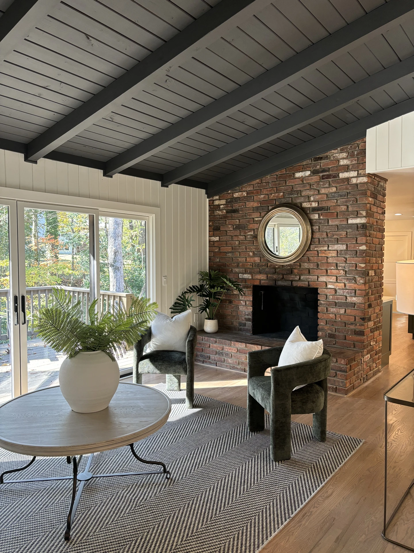 Bright living room makeover featuring a rustic brick fireplace, dark wood beam ceiling, and large sliding doors letting in natural light. Two modern green chairs with white cushions sit on a fresh herringbone-patterned rug, showing how easily you can transform a space when you replace your rug. A round wooden coffee table with a white vase of greenery adds an organic, welcoming touch.