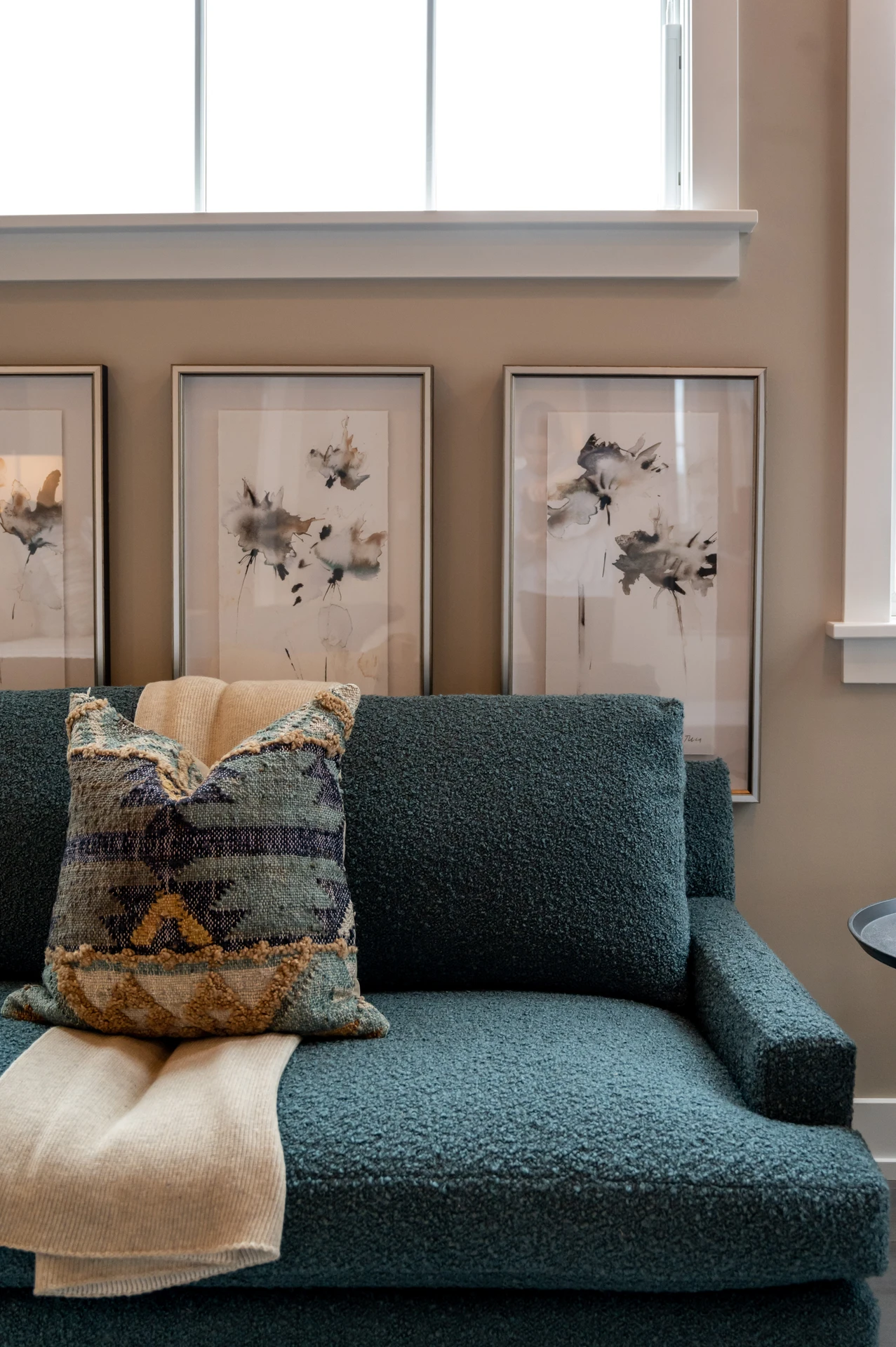 A modern living room makeover featuring a textured teal sofa accented with a patterned pillow and a light knit throw draped casually for warmth. Behind the seating, three abstract floral prints in muted tones line the wall, adding softness and elegance. The setup highlights how cozy throws and blankets can instantly make a seating area more inviting while tying the colors of the decor together.