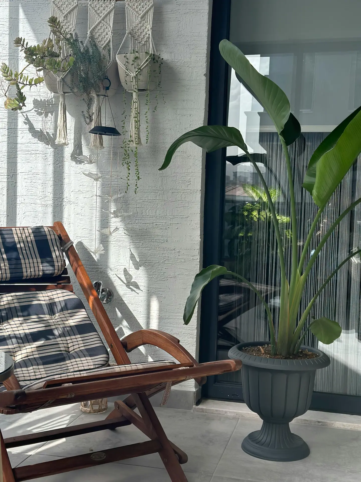 A cozy balcony styled with a wooden deck chair featuring plaid cushions, complemented by macrame plant hangers on the wall and a tall green plant in a decorative pot. The mix of natural textures and greenery provides inviting inspiration for balcony decoration ideas that blend comfort with boho charm.