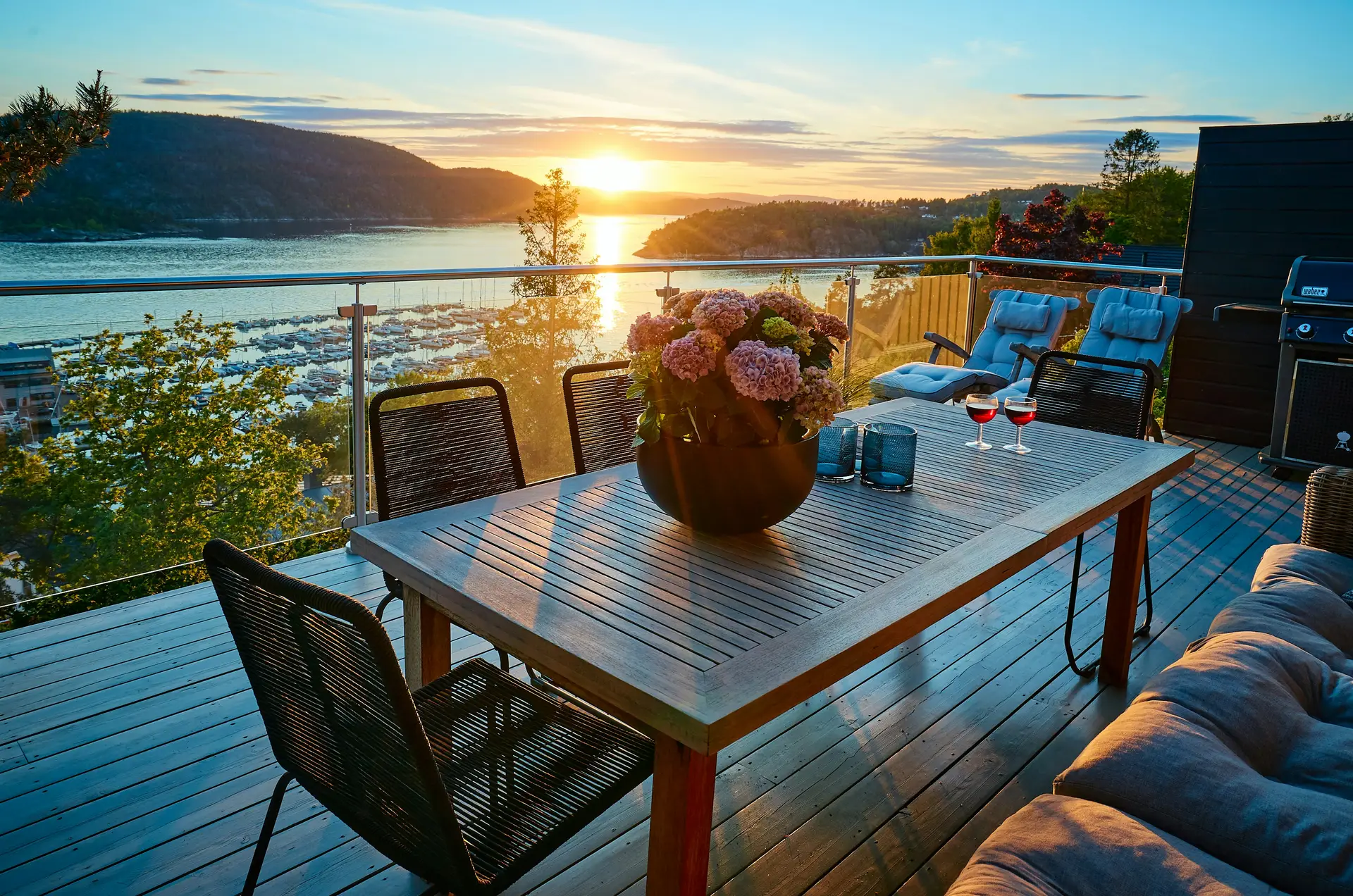 Modern terrace overlooking a marina and mountains at sunset, styled with a wooden dining table centerpiece, black chairs, and soft lounge seating. A large flower arrangement and candles add charm, making this a perfect setting for relaxing evenings and inspiring balcony decoration ideas for combining comfort with scenic views.