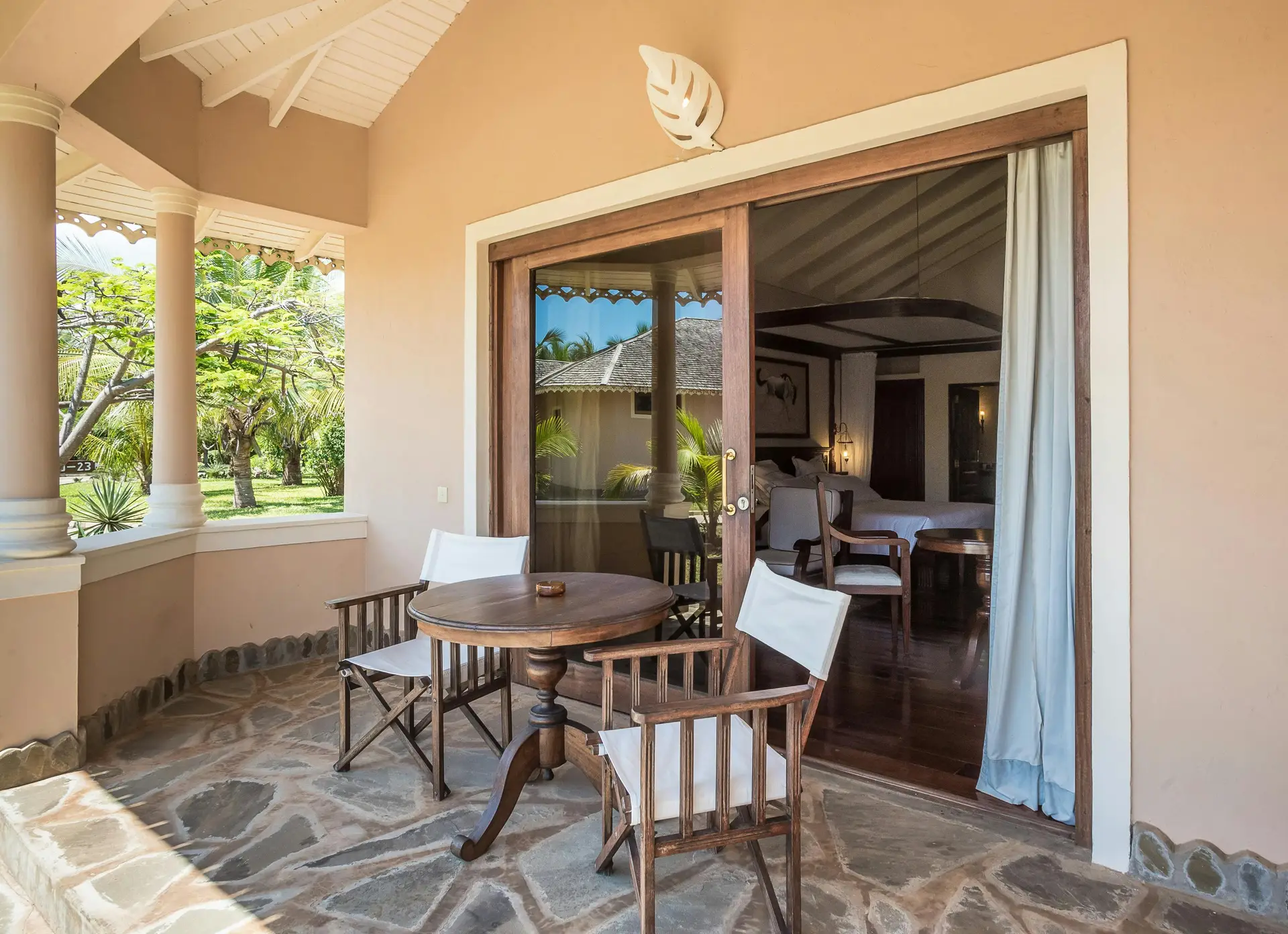 Covered terrace with rustic stone flooring, a small wooden round table, and two classic director's chairs, overlooking a lush green garden. A charming mix of comfort and simplicity, this setup offers inspiration for balcony decoration ideas that embrace natural materials and cozy outdoor living.