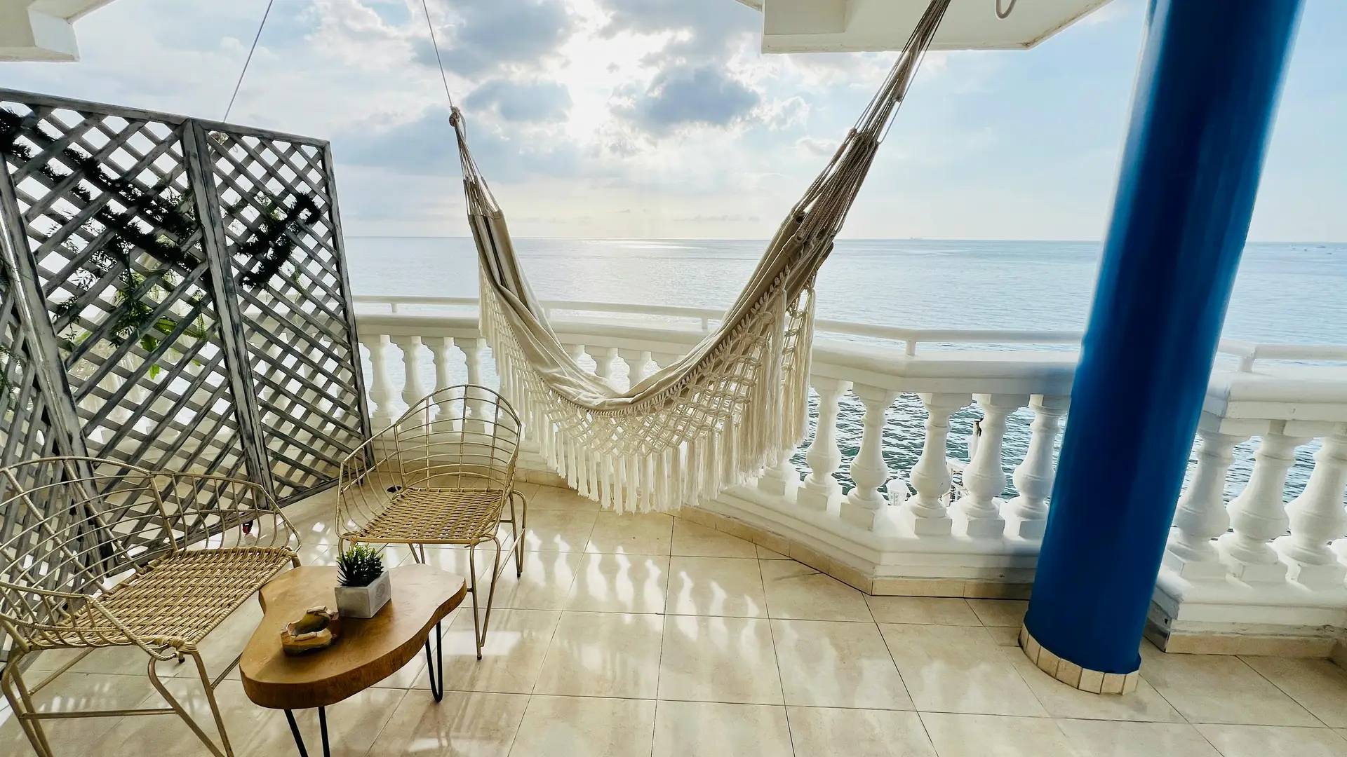 Seaside balcony featuring a cozy hammock draped in macramé style, a pair of woven metal chairs, and a small wooden coffee table with plants, offering serene ocean views. A perfect showcase of balcony decoration ideas that mix comfort, style, and relaxation.