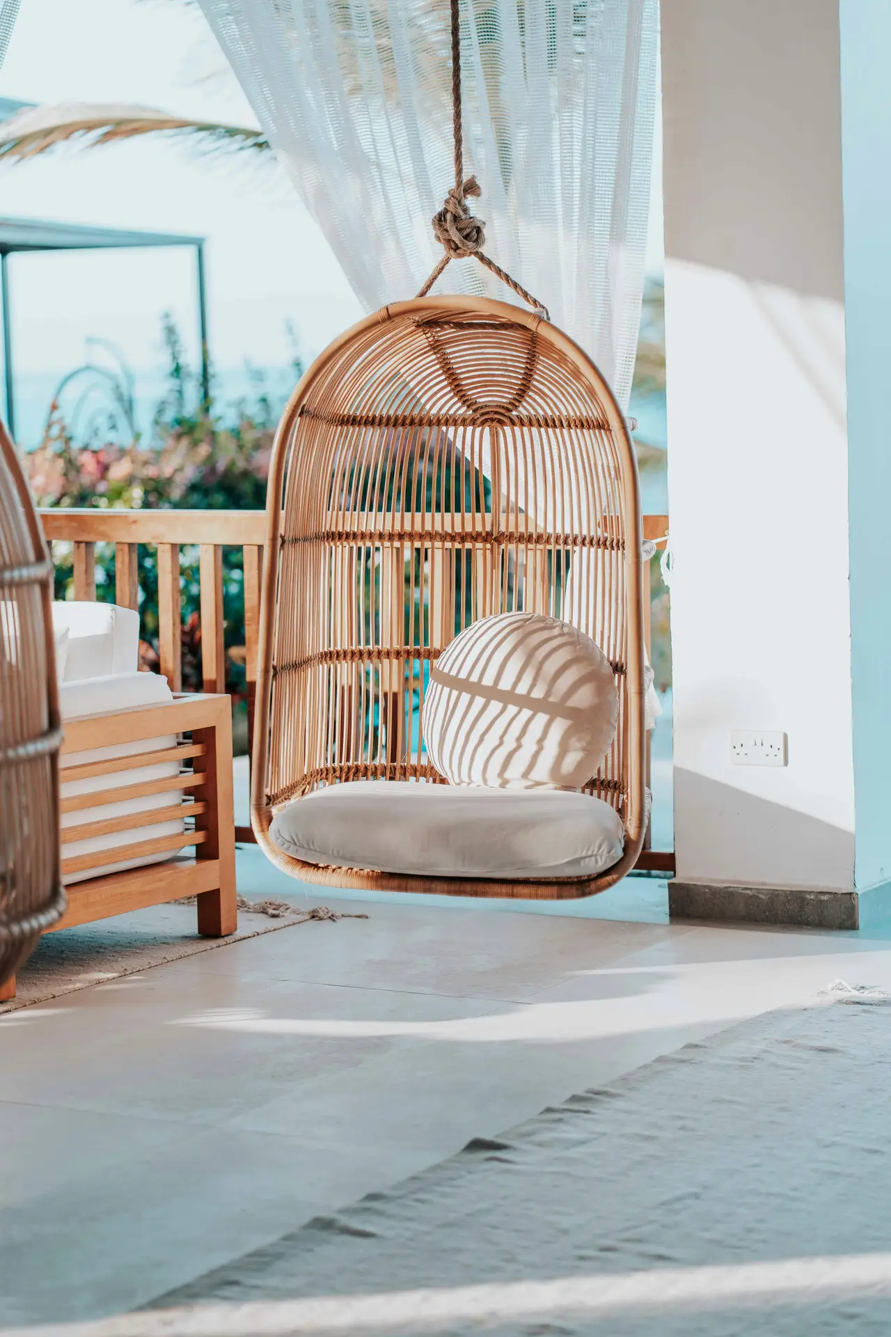 A sunlit balcony styled with a woven hanging chair featuring a soft cushion and round pillow, paired with light curtains that let natural light filter through. This cozy setup highlights modern balcony decoration ideas, blending comfort and relaxation with airy, coastal-inspired vibes.
