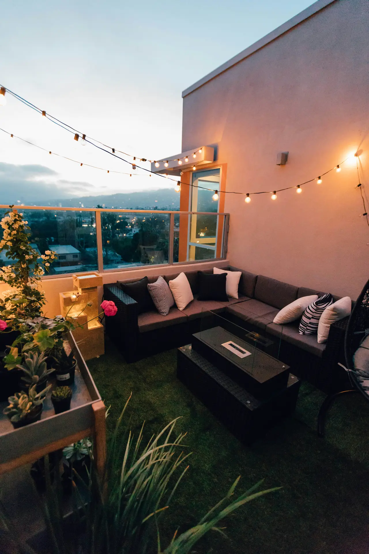 A cozy evening balcony setup featuring an L-sectional sofa with patterned and neutral cushions, string lights hanging overhead, a sleek firepit-style table, and potted plants adding greenery. Perfect for modern balcony decoration ideas that mix comfort, style, and ambiance for entertaining or unwinding.