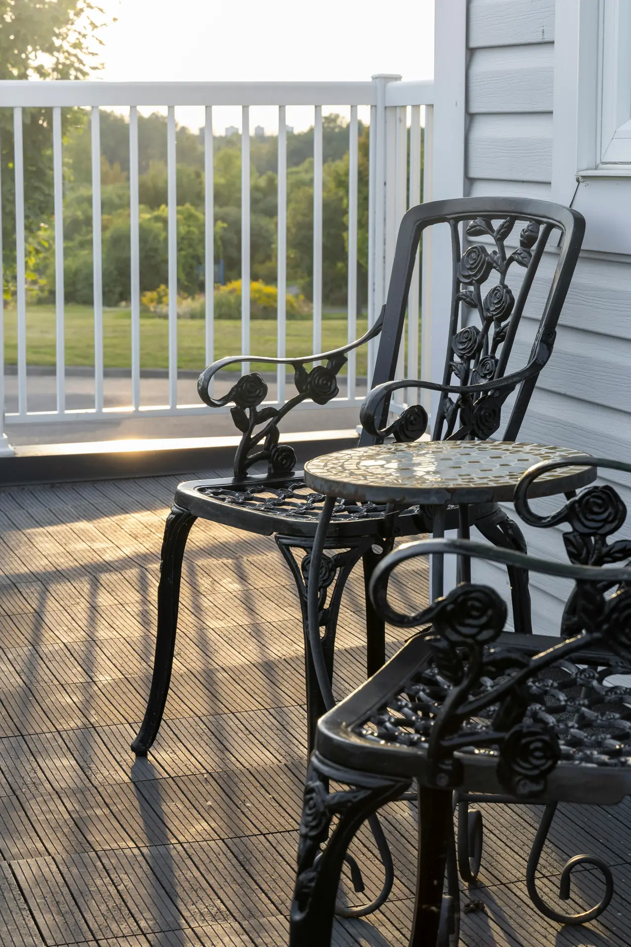 Elegant metal decorated chairs with floral detailing paired with a small mosaic-topped round table on a cozy balcony, overlooking a lush green landscape at sunset. A perfect example of balcony decoration ideas that blend durability with charm, creating an inviting nook for enjoying morning coffee or evening relaxation.