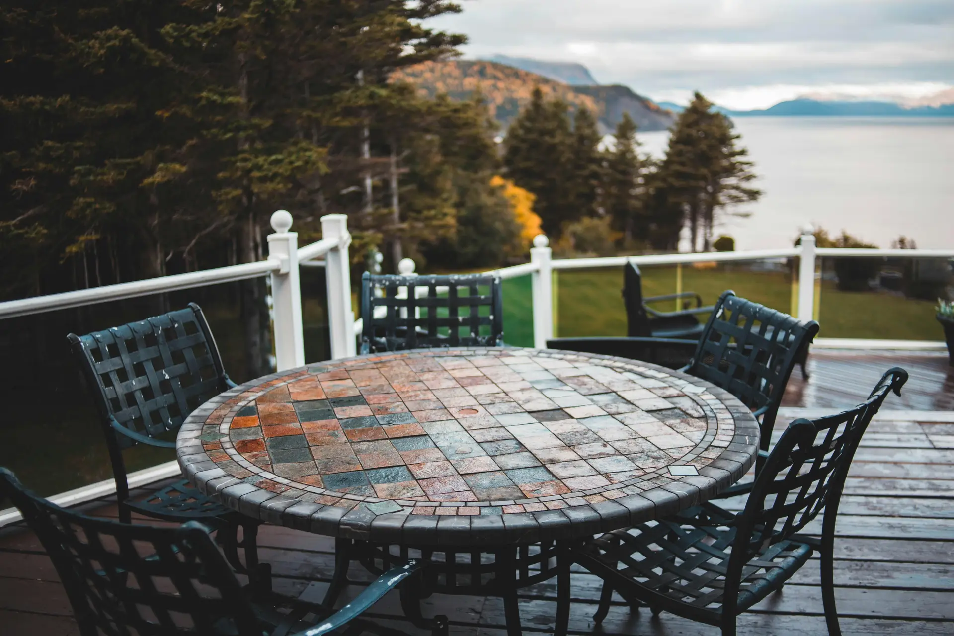 Scenic balcony with a mosaic decorated round table surrounded by dark woven metal chairs, overlooking a stunning lake and forested mountains. A great example of rustic yet stylish balcony decoration ideas, blending outdoor dining comfort with a natural panoramic view.