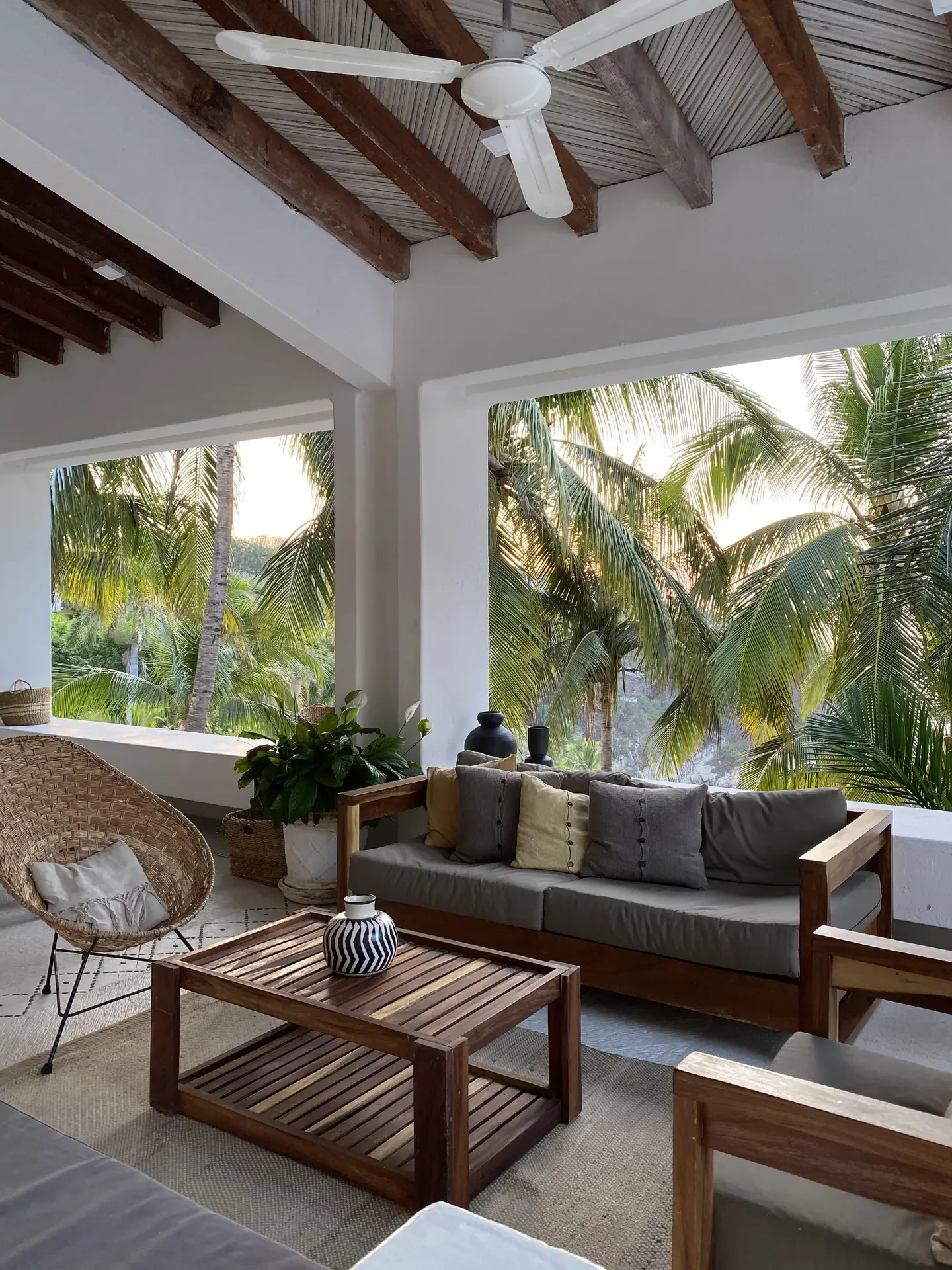 Stylish tropical-inspired outdoor seating area that showcases creative balcony decoration ideas. Wooden furniture with neutral cushions, a woven chair, and a slatted coffee table add warmth, while potted plants bring freshness. The ceiling fan, exposed beams, and open views of lush palm trees create a breezy, resort-like vibe perfect for relaxation or entertaining.