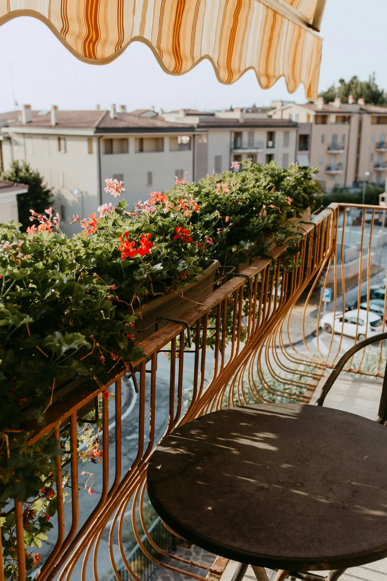 A charming urban balcony with golden railings, adorned with lush geraniums in plant boxes for the railing, creating a burst of color above a small round table. The striped awning adds shade, making this a cozy spot and offering inspiration for simple yet vibrant balcony decoration ideas.