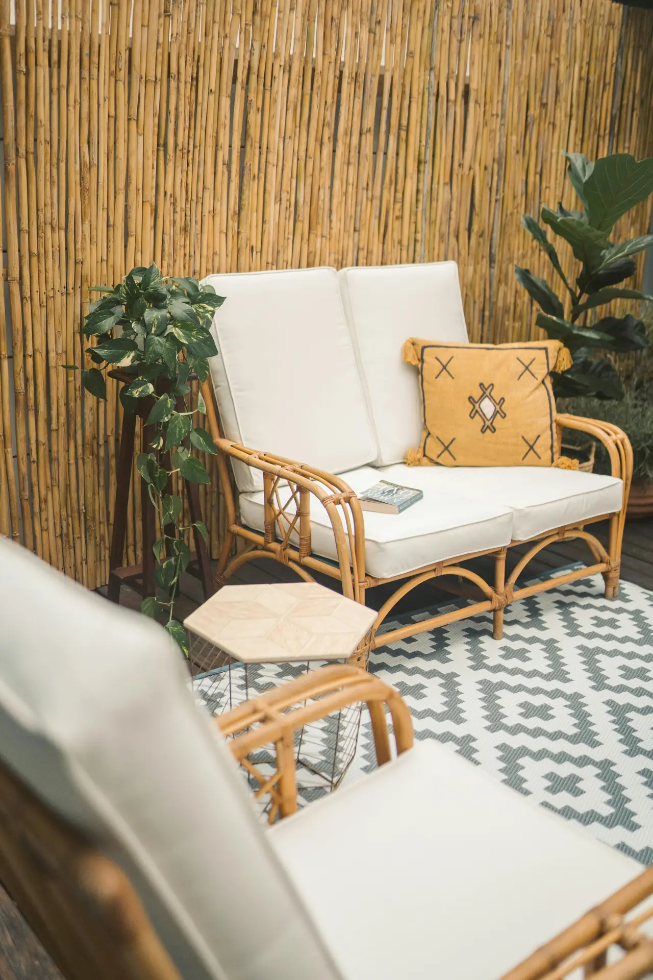 A cozy balcony corner featuring small rattan sofas with white upholstery, styled with a patterned rug, green plants, and a bamboo privacy wall. A mustard cushion adds a pop of color, showcasing modern yet natural balcony decoration ideas for a relaxed and stylish outdoor retreat.