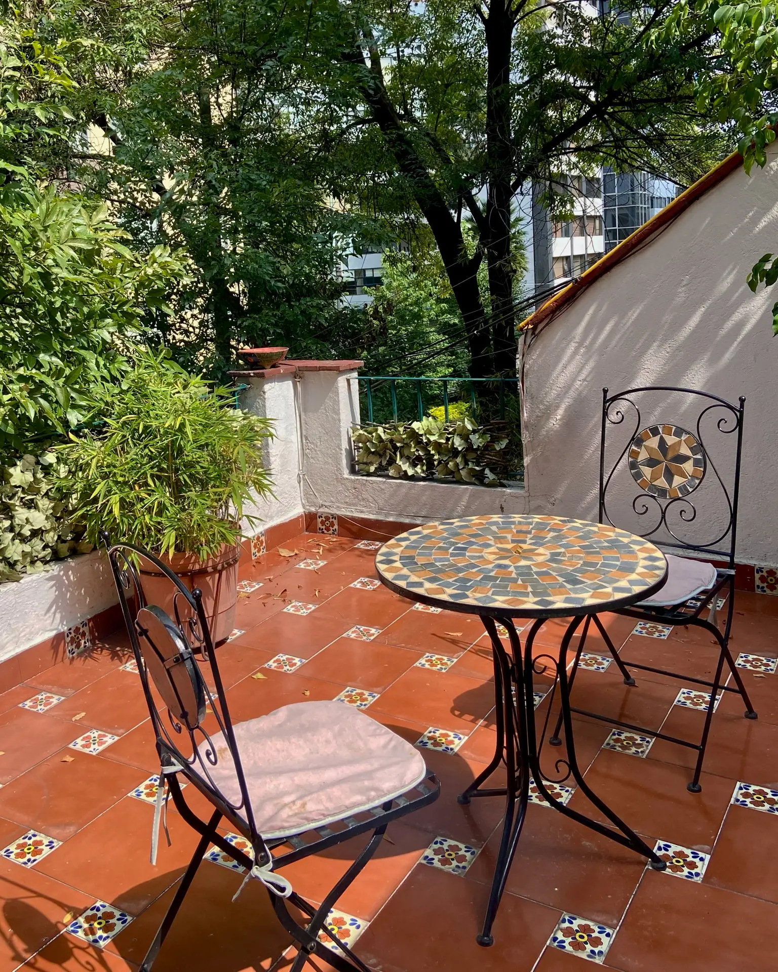 Cozy outdoor nook showcasing balcony decoration ideas with a terracotta-tiled floor accented by colorful patterns. A small table with small chairs featuring mosaic designs creates a charming setup for morning coffee or evening relaxation. Surrounded by potted greenery and shaded by trees, this intimate balcony blends natural elements with artistic details for a welcoming vibe.